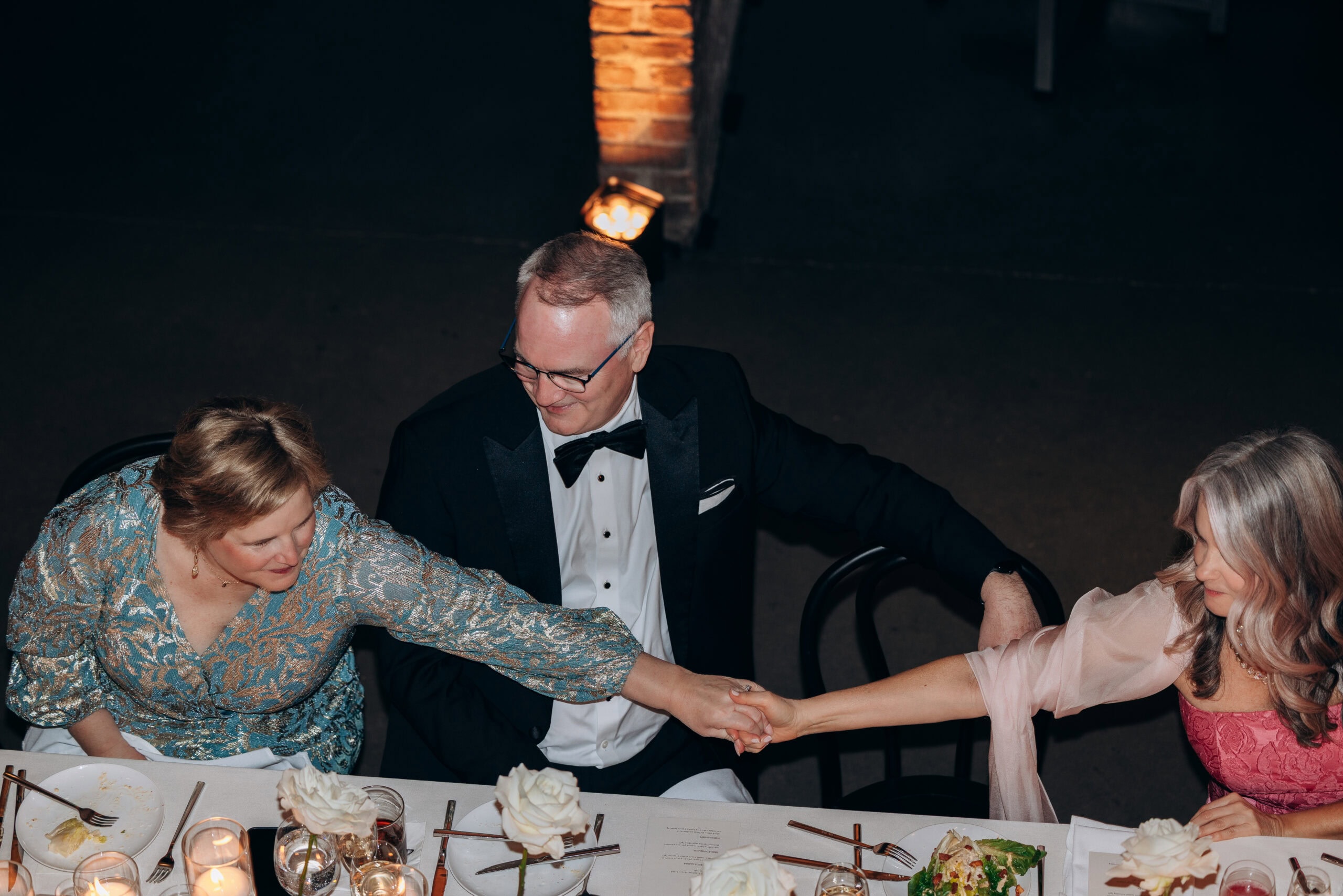 Three formally dressed wedding guests seated at a candlelit reception table at The Foundry, reaching across the table to hold hands.