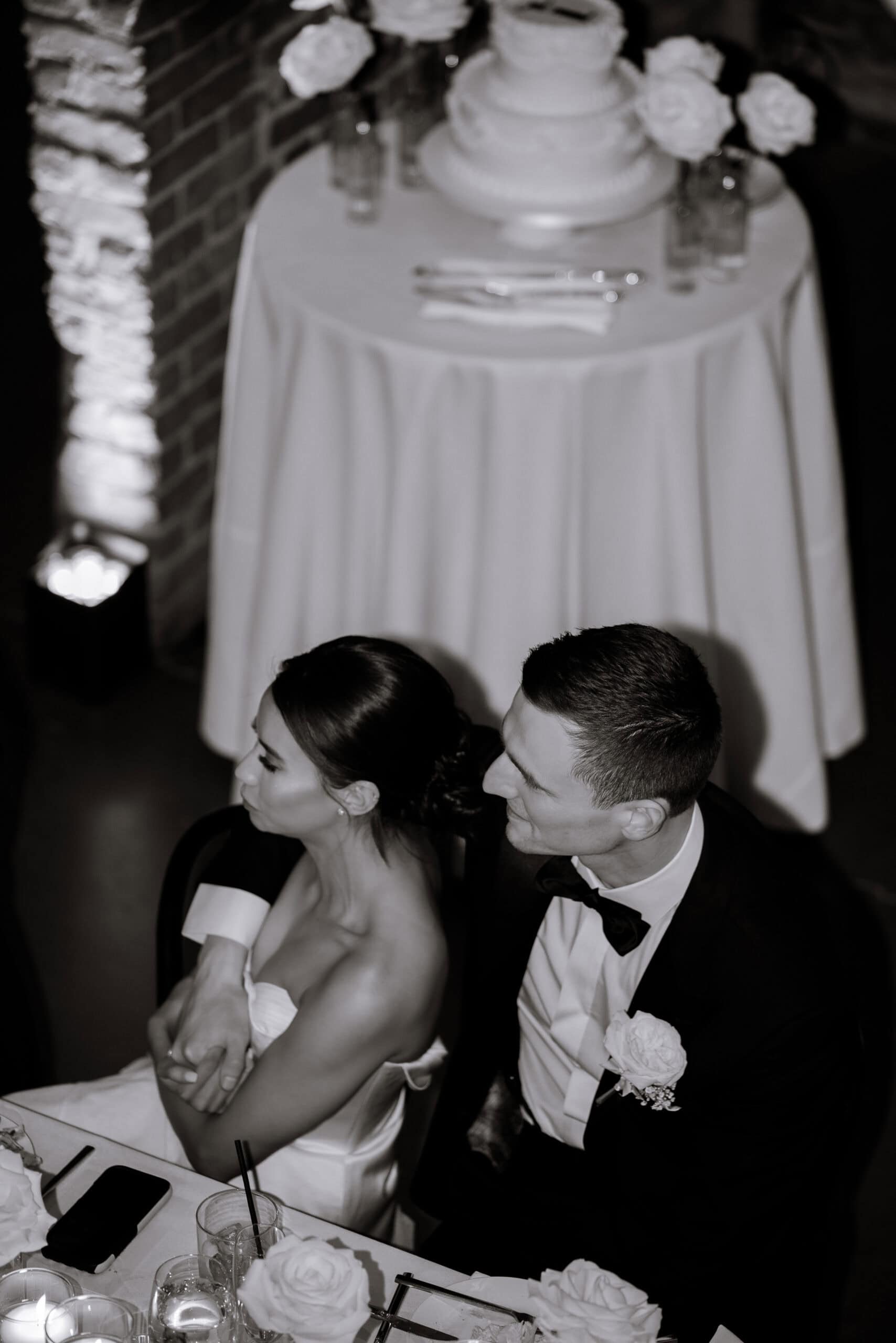 Overhead black-and-white photo of a seated couple at their wedding reception at The Foundry, with a tiered cake, white roses, candles, and drinks on surrounding tables.