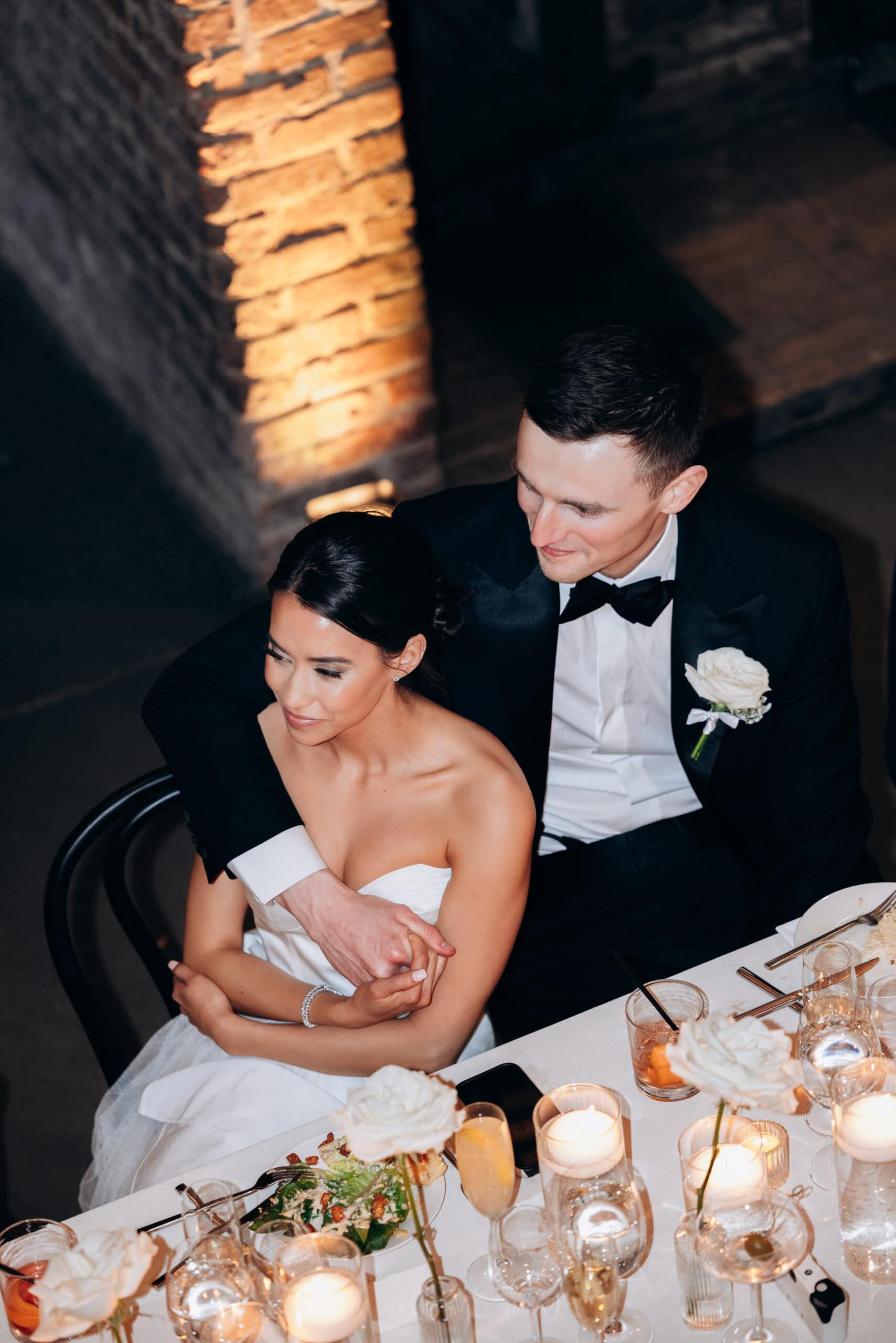 Overhead view of a bride in a strapless gown and a groom in a black tuxedo seated closely at a candlelit reception table with white roses at The Foundry.