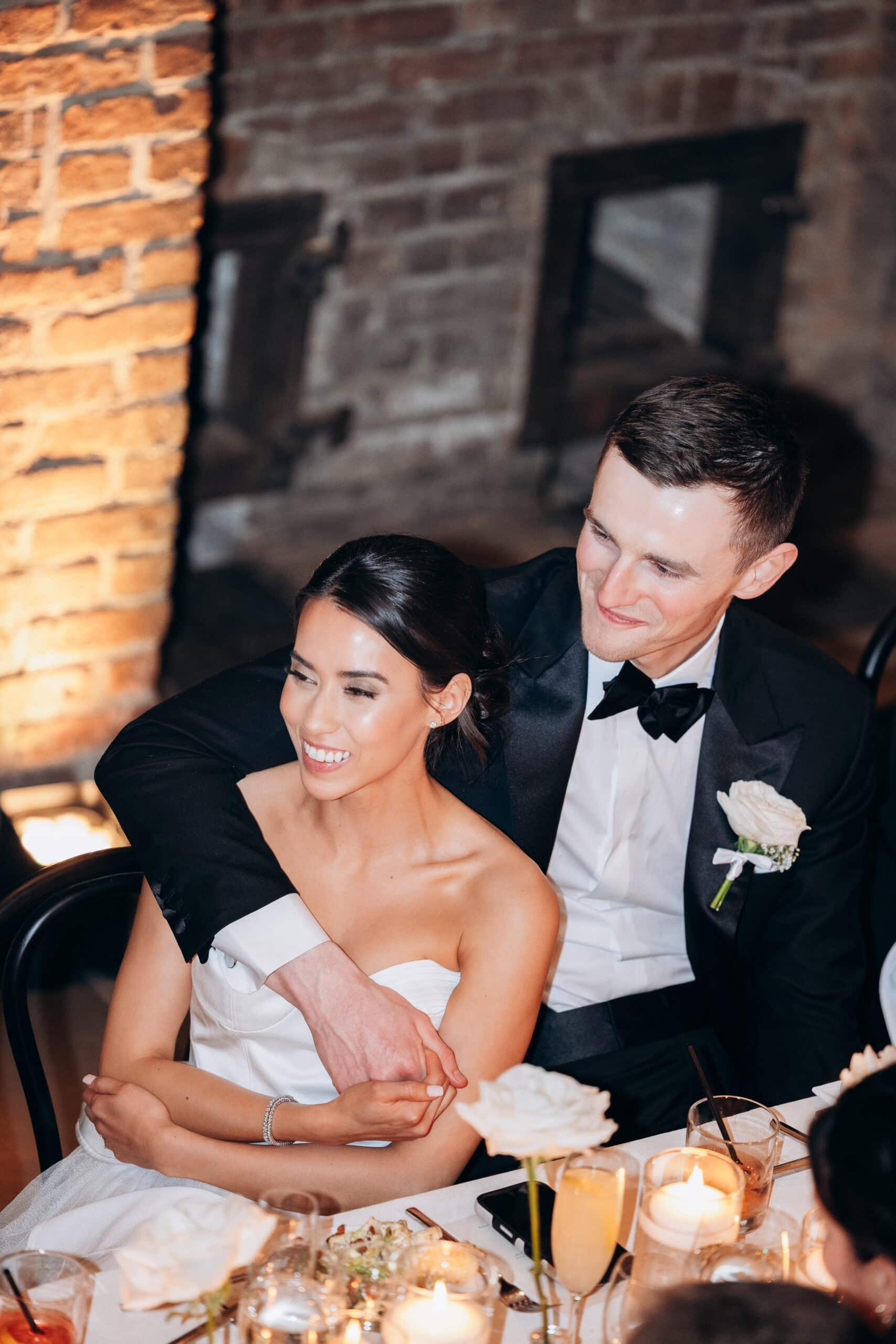 Bride in a strapless white gown seated at a candlelit reception table as the groom in a black tuxedo embraces her from behind at The Foundry.