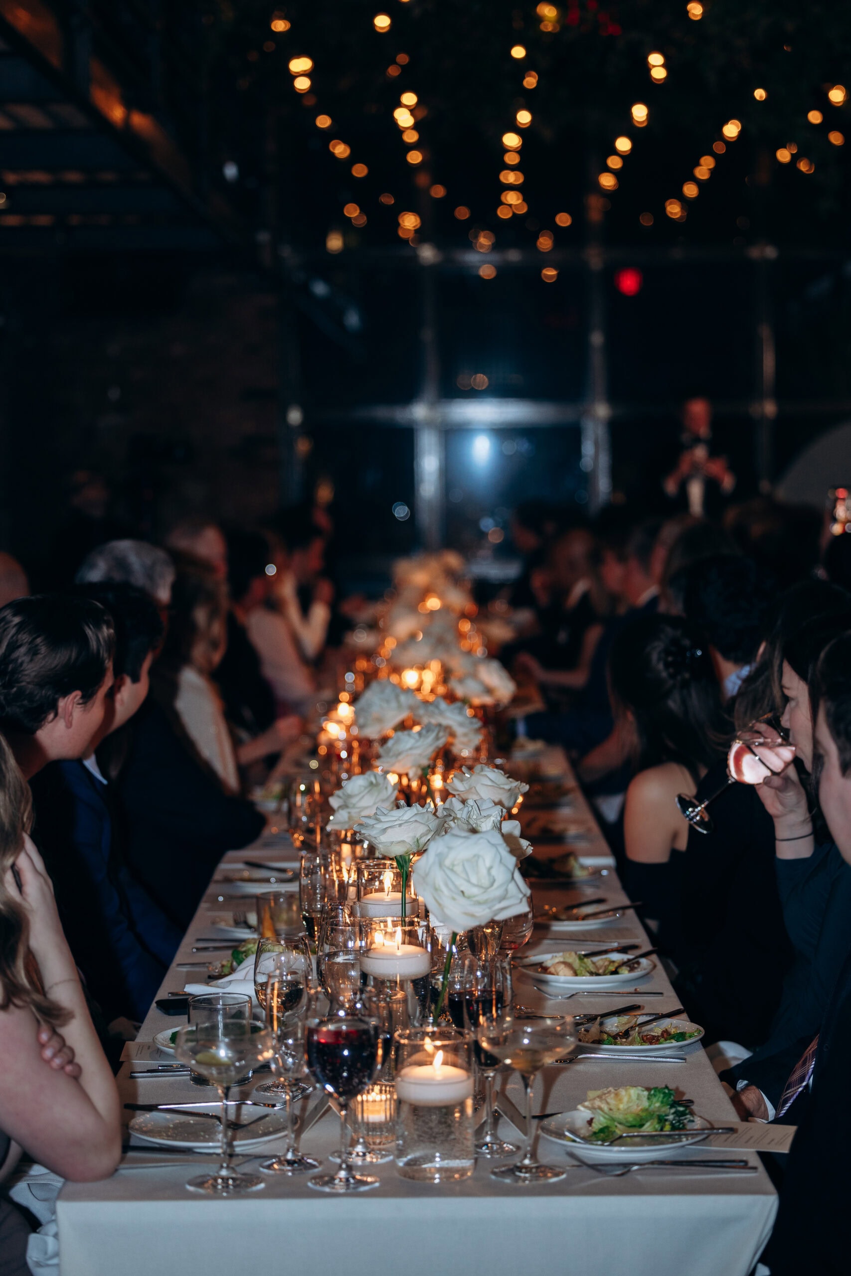 Guests seated at a long candlelit wedding reception table with white rose centerpieces and string lights overhead at The Foundry.