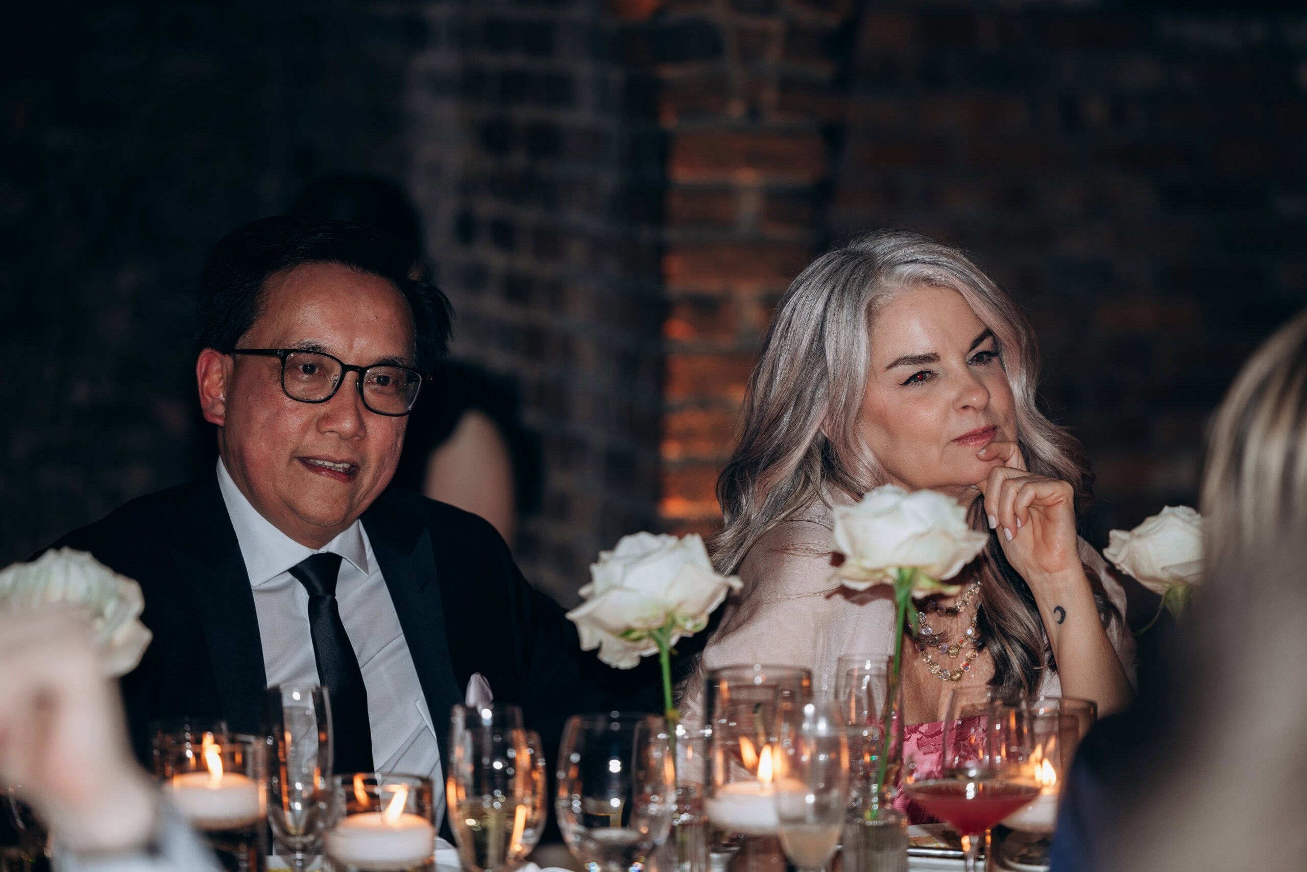 Two wedding guests seated at a candlelit reception table at The Foundry, with white roses, glassware, and exposed brick in the background.