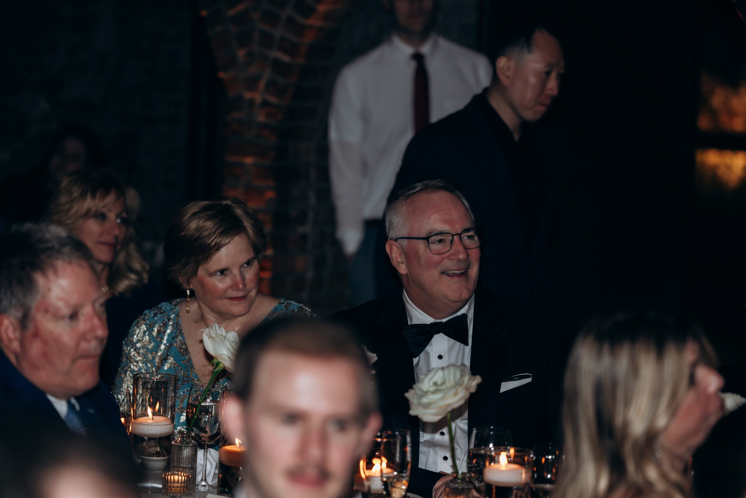 Wedding guests seated at a candlelit reception table inside The Foundry, with white roses, glassware, and brick arches in a dimly lit interior.