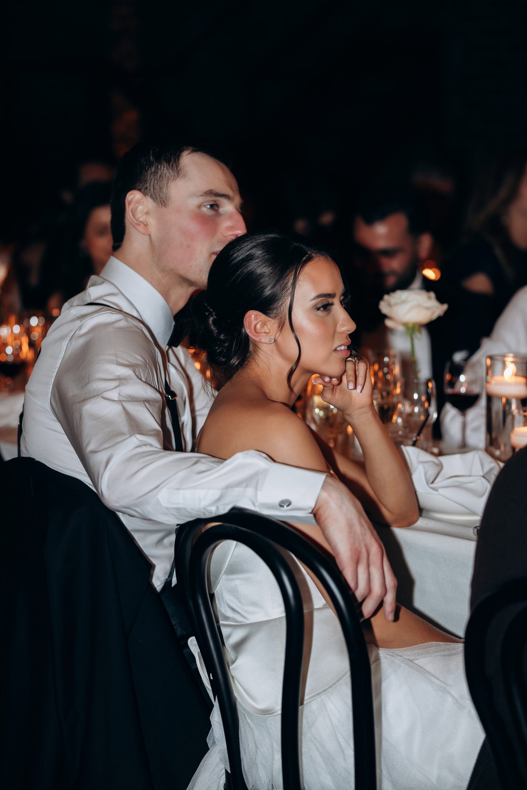 Bride and groom seated closely at a candlelit reception table at The Foundry, with wine glasses, a white rose, and dark ambient background.
