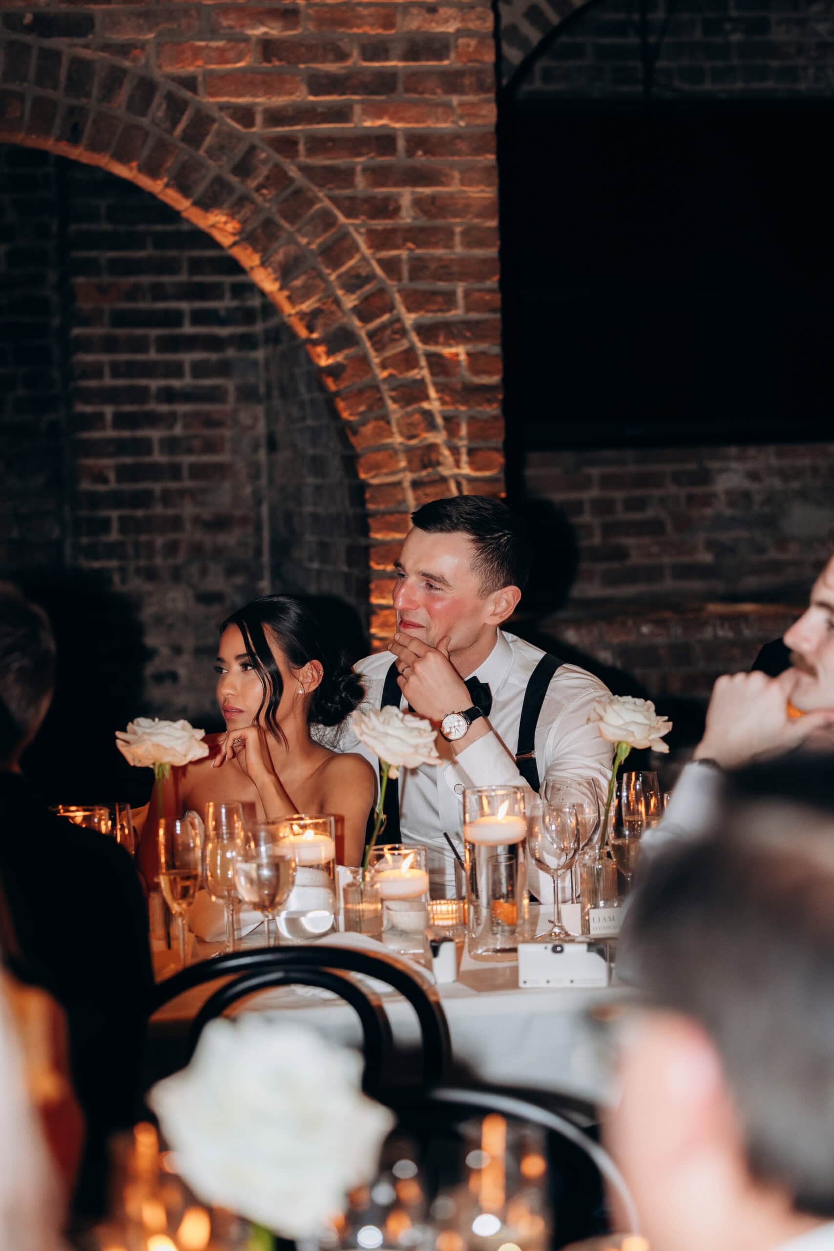 Bride and groom seated at a candlelit reception table at The Foundry, surrounded by white roses, glassware, and dramatic brick arches.