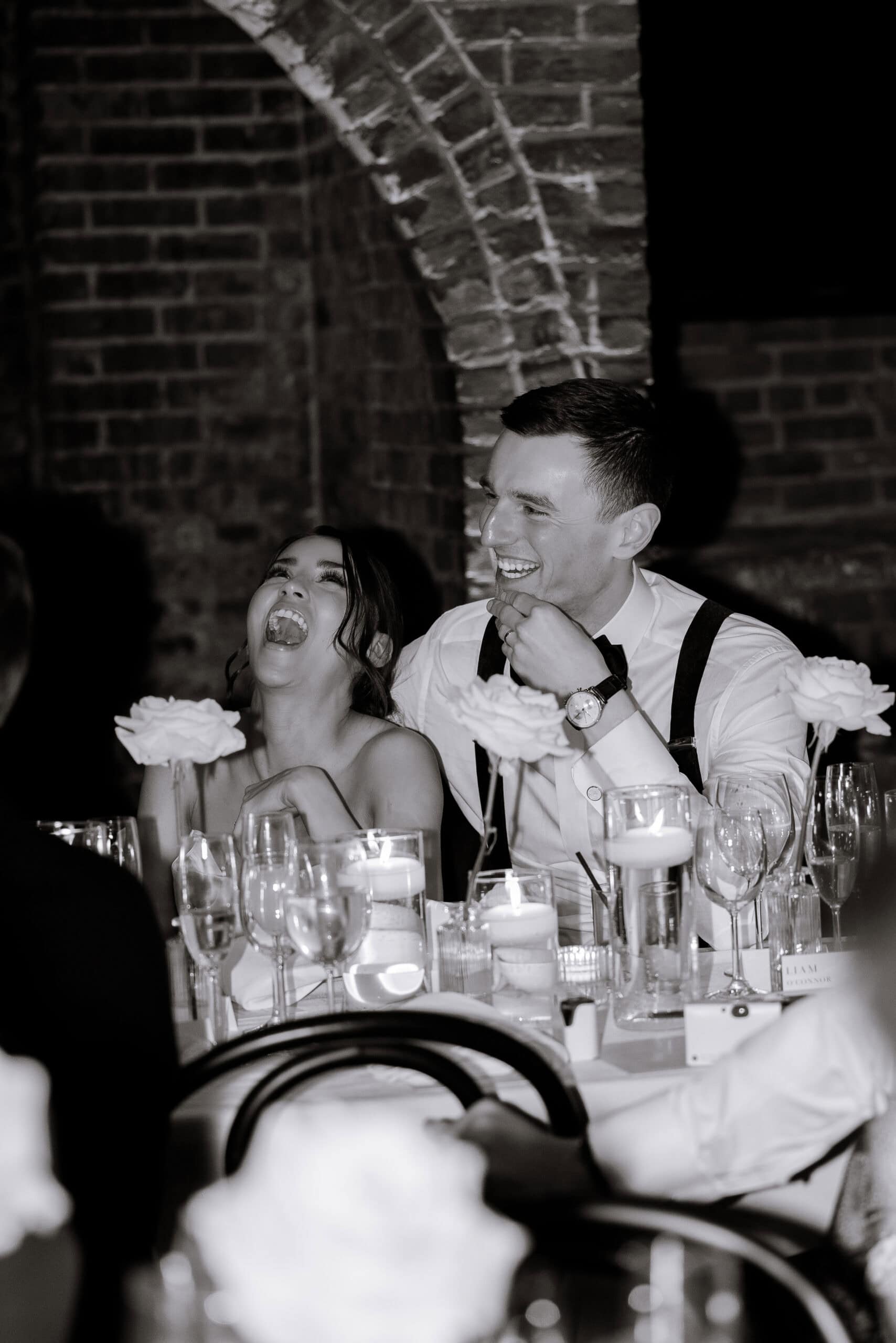 Black-and-white wedding reception scene at The Foundry with a couple laughing at a candlelit table beneath brick arches and white floral arrangements.