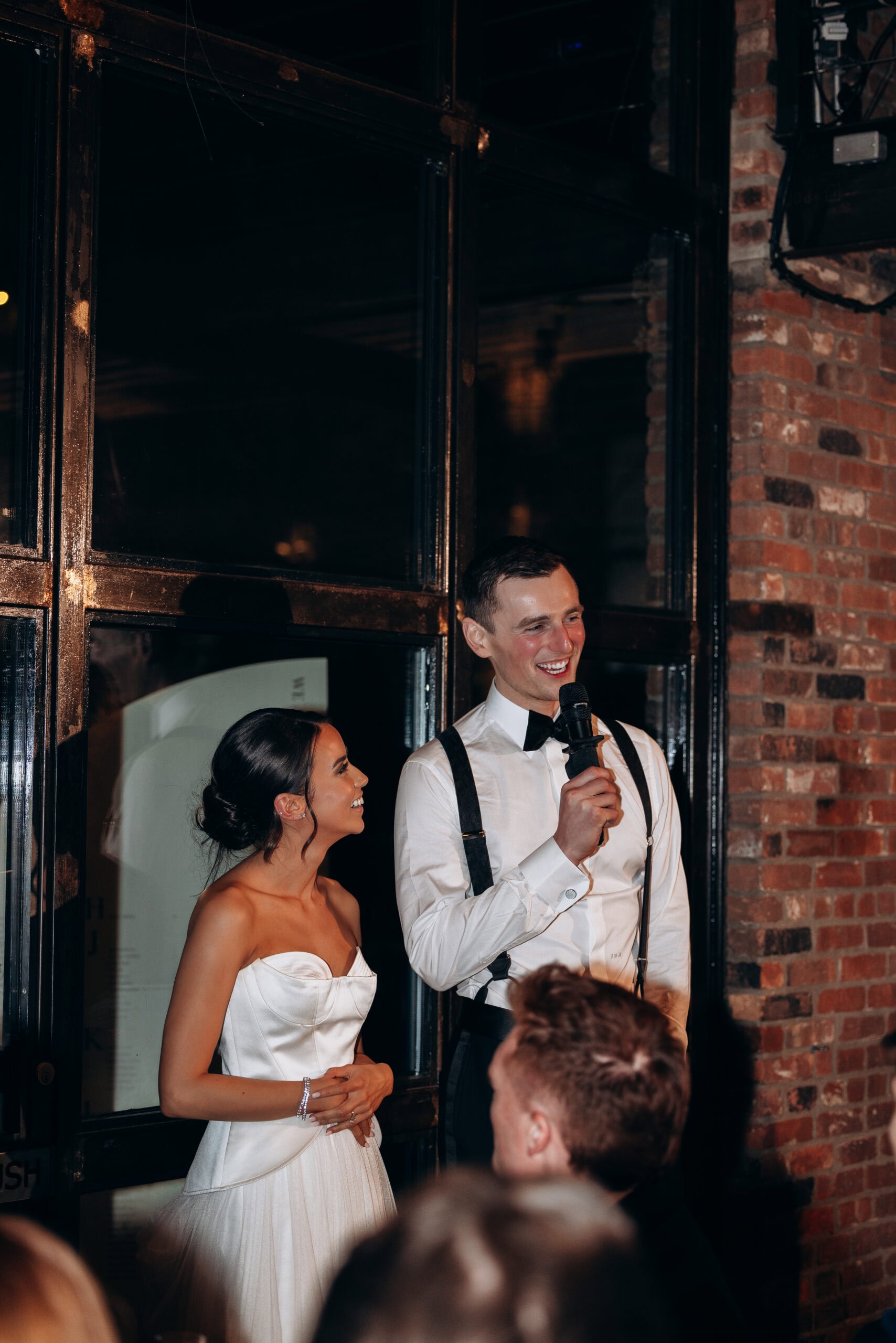 Bride and groom standing indoors at The Foundry during the reception, as the groom gives a speech into a microphone beside exposed brick and black-framed windows.