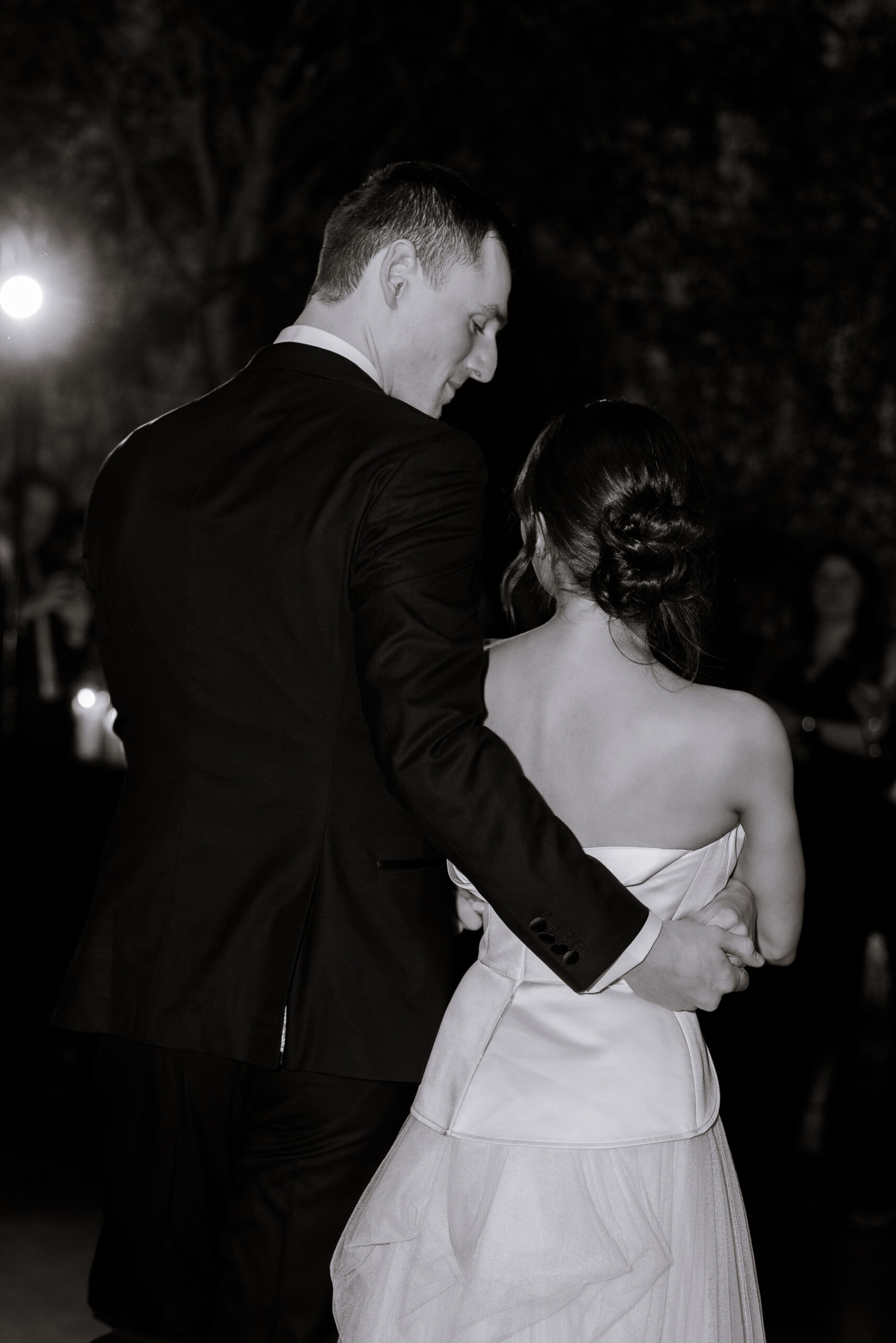 Black-and-white photo of a couple from behind, embracing during an evening wedding reception at The Foundry, with soft lights and blurred guests in the background.