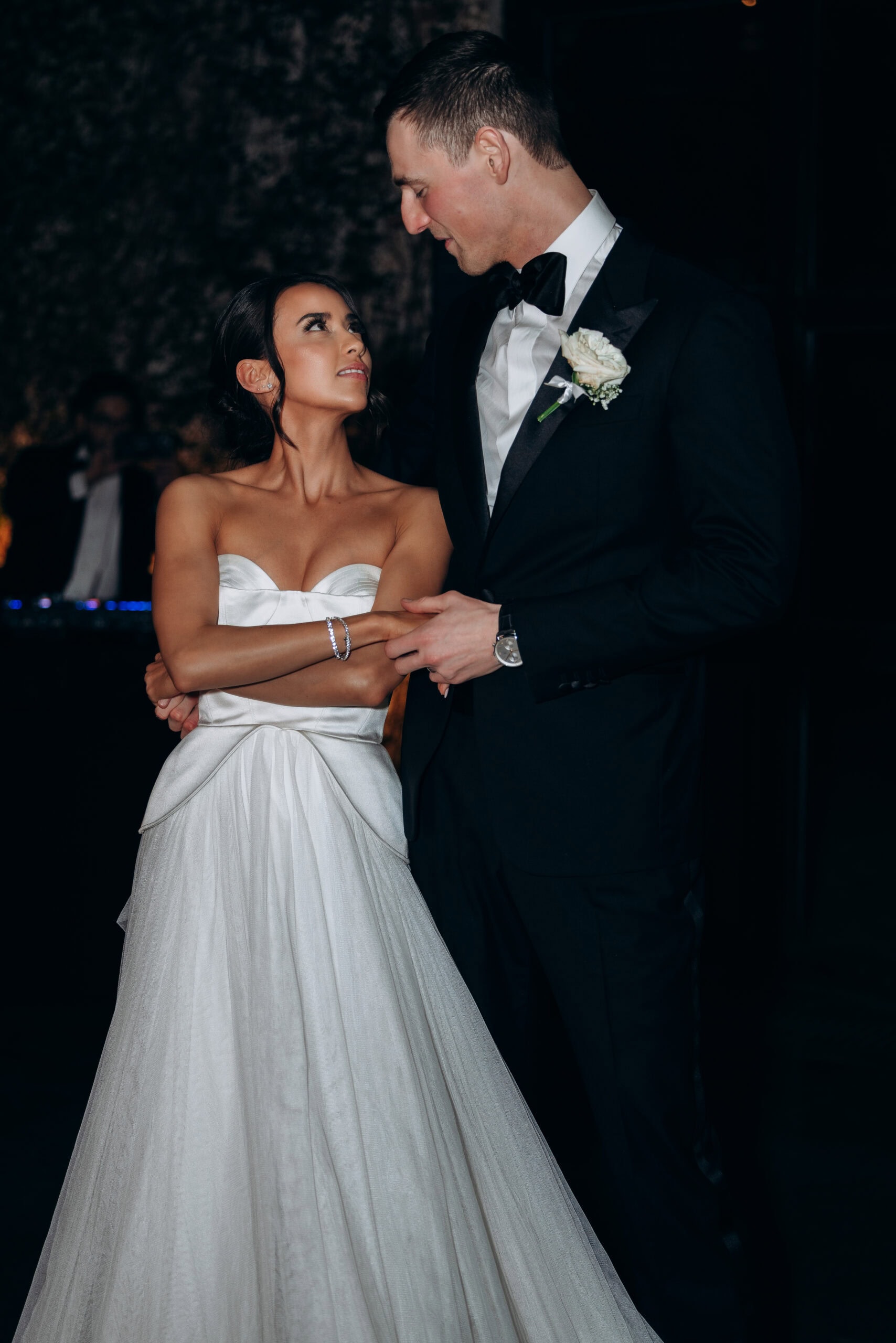Bride in a strapless satin wedding gown stands beside groom in a black tuxedo at The Foundry, sharing an intimate look in a dimly lit reception space.
