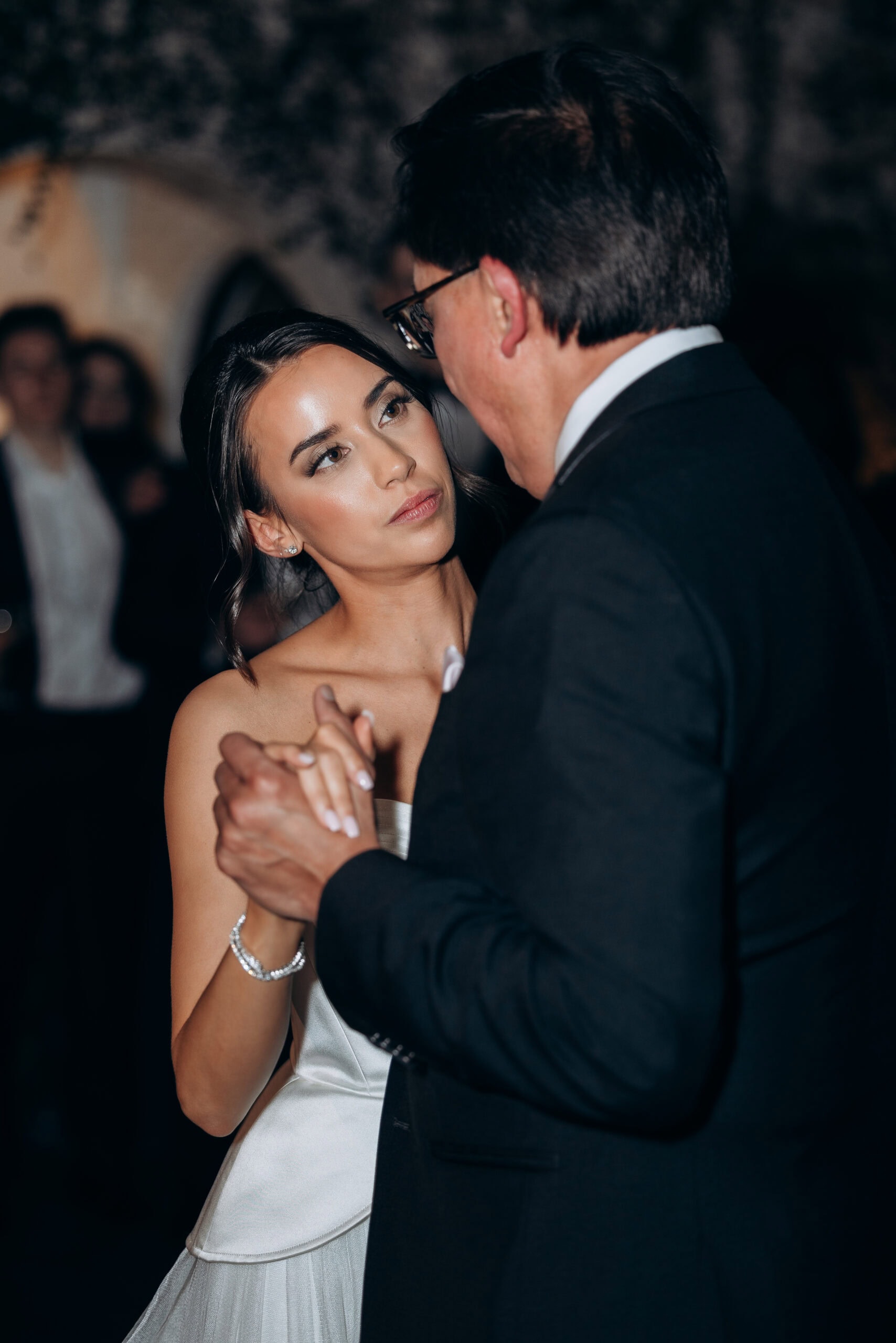 Bride in a satin wedding gown slow dances with an older man in a dark suit during a reception at The Foundry, with guests softly blurred in the background.