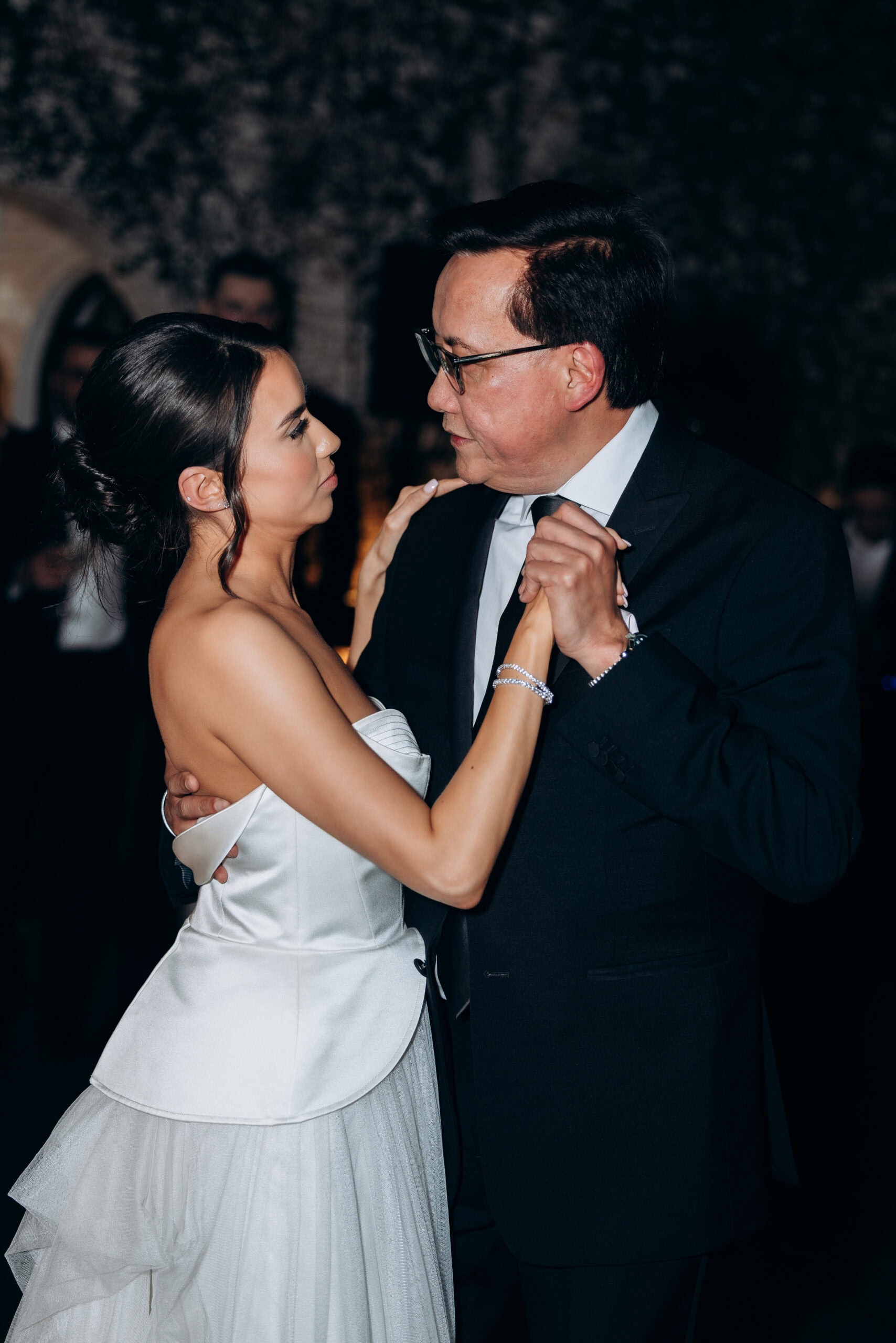 Bride in a structured satin wedding gown dances closely with an older man in a black suit during the reception at The Foundry, in a dimly lit interior.