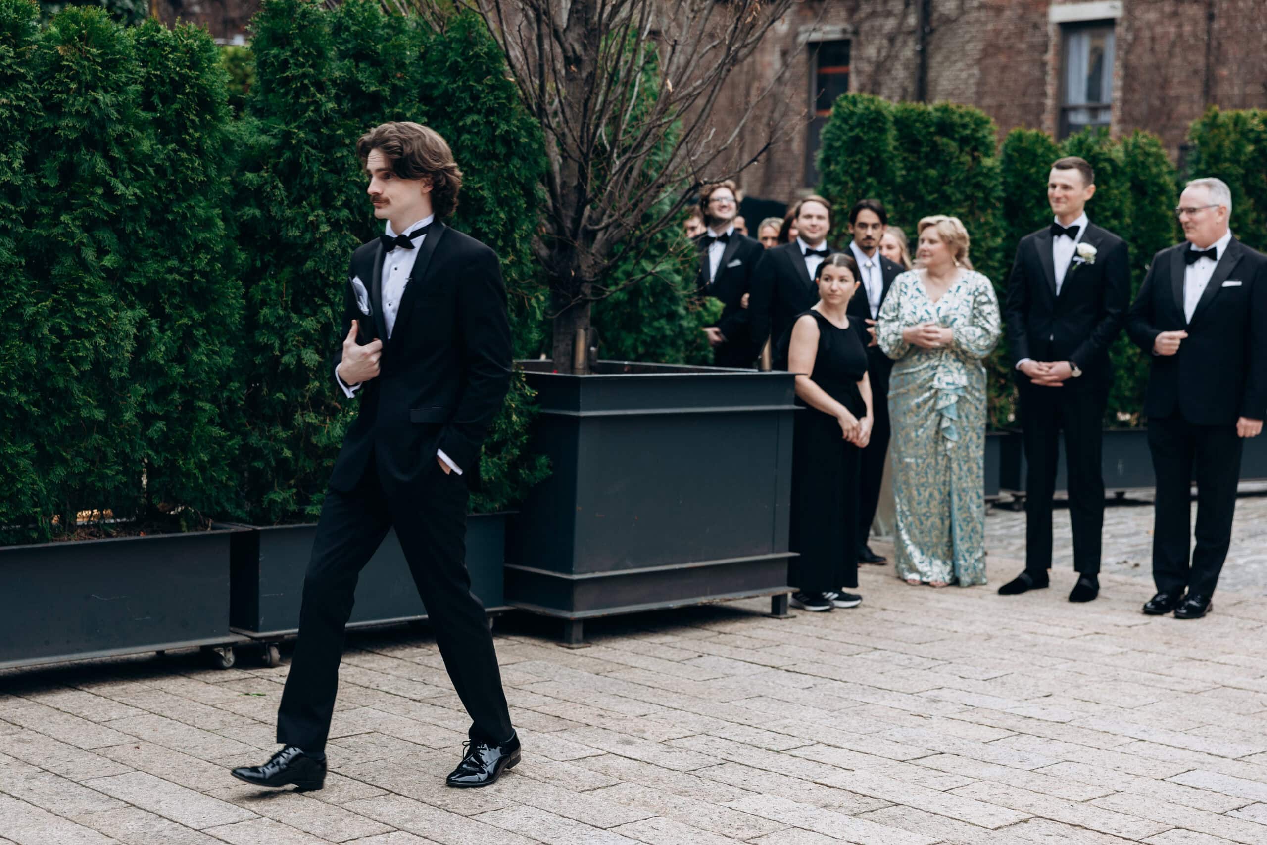A man in a black tuxedo walks across The Foundry courtyard as formally dressed wedding guests stand beside large planters and hedges.