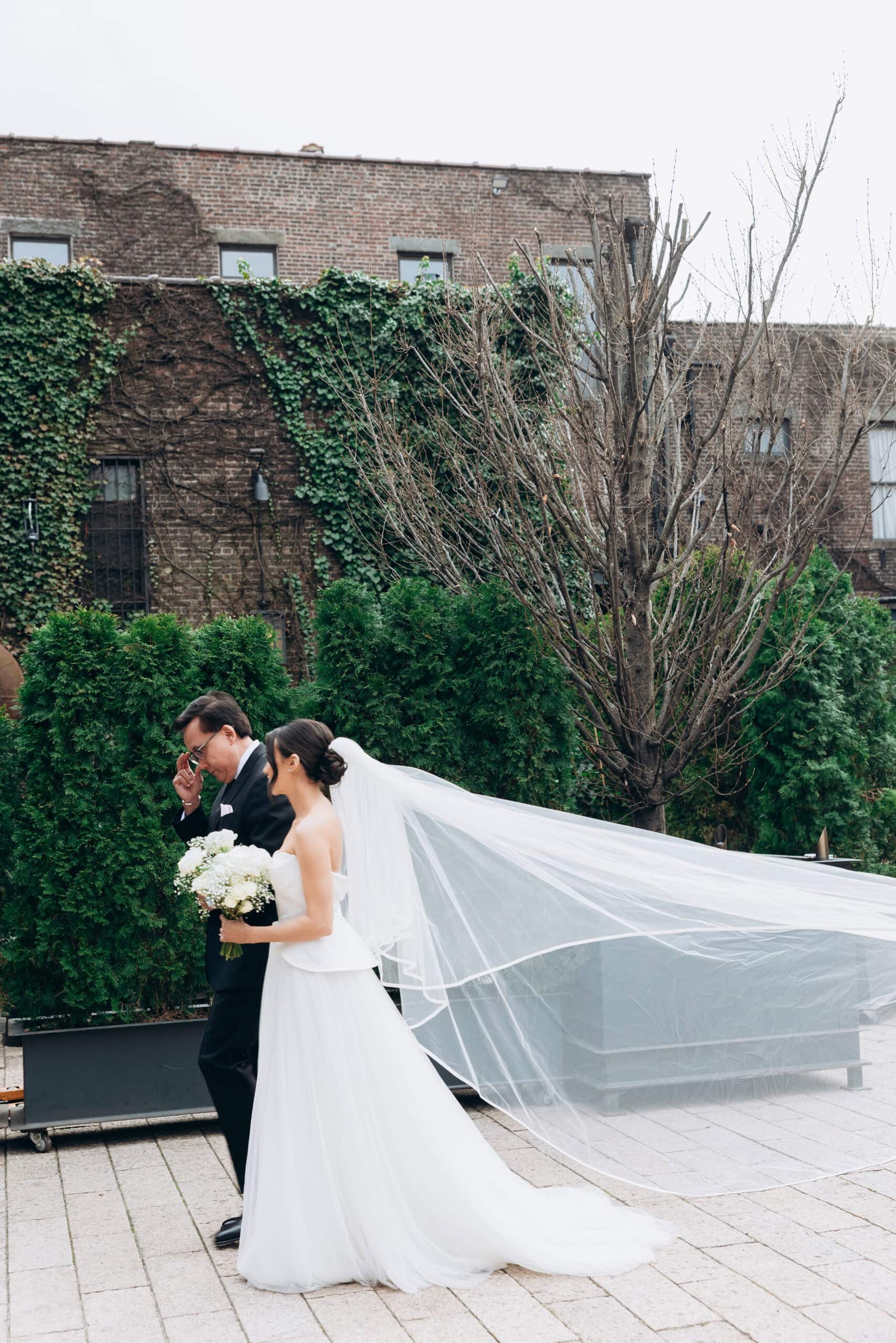 Bride in a strapless gown with a long flowing veil walks beside a man in a black suit through the courtyard at The Foundry, framed by ivy-covered brick walls.