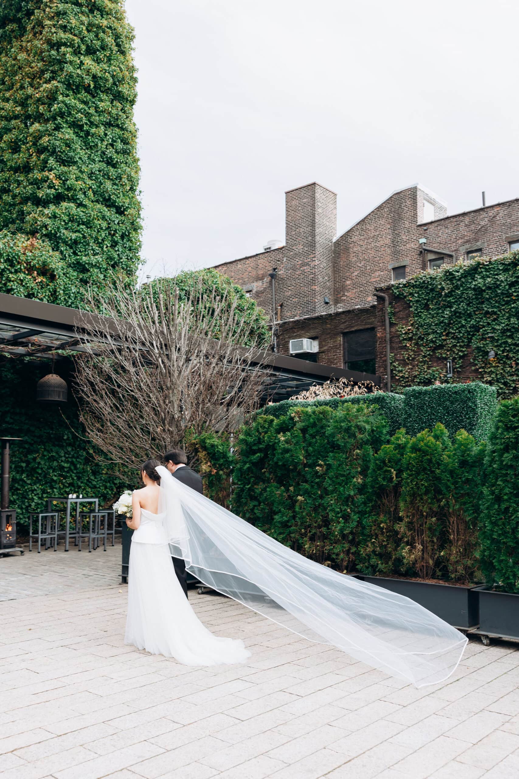Bride and partner walk through The Foundry courtyard as her long veil streams behind her, framed by ivy-covered brick walls, hedges, and pale stone paving.