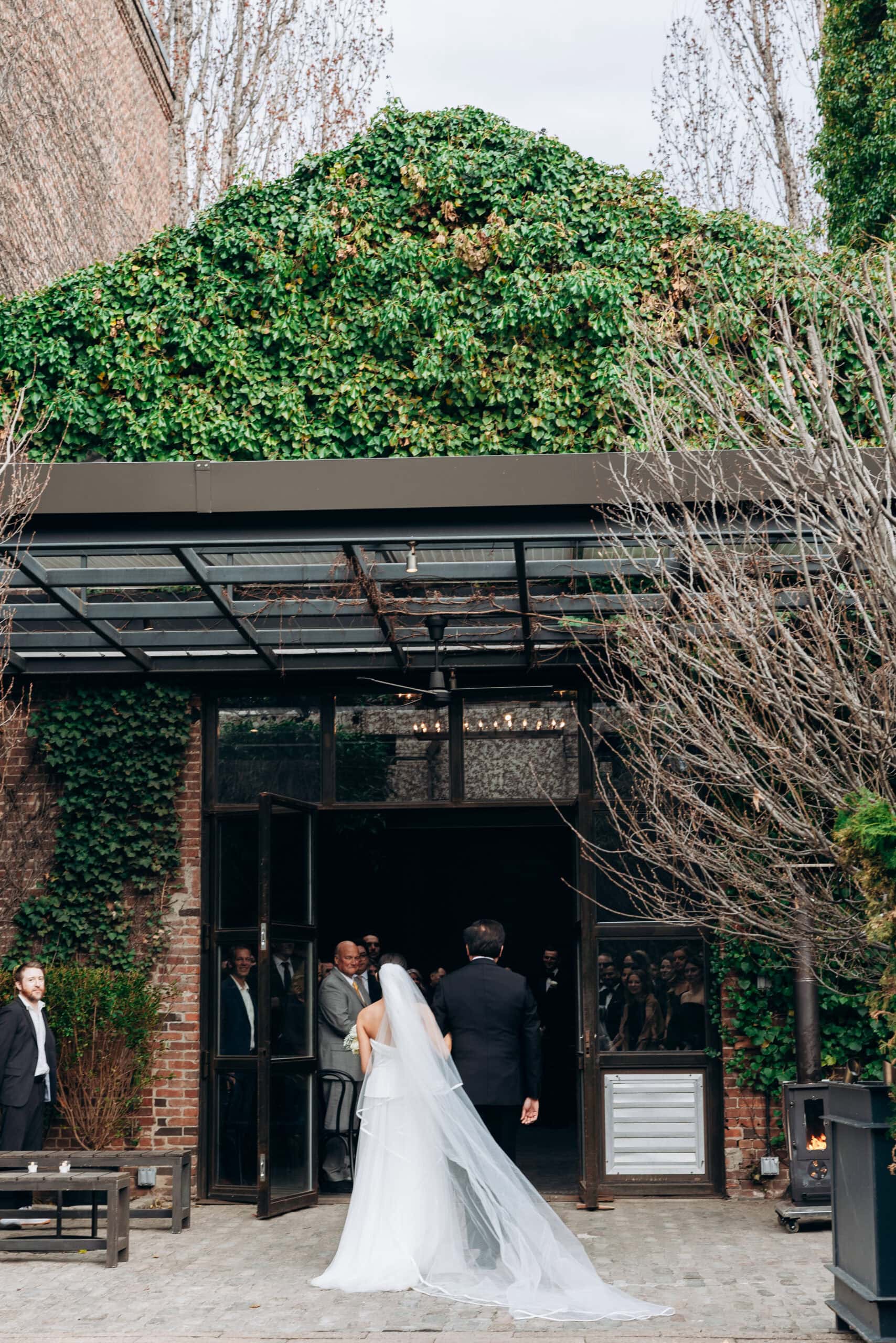 Bride in a strapless gown and long veil walks into The Foundry with a suited escort, framed by ivy-covered brick, glass doors, and gathered guests.