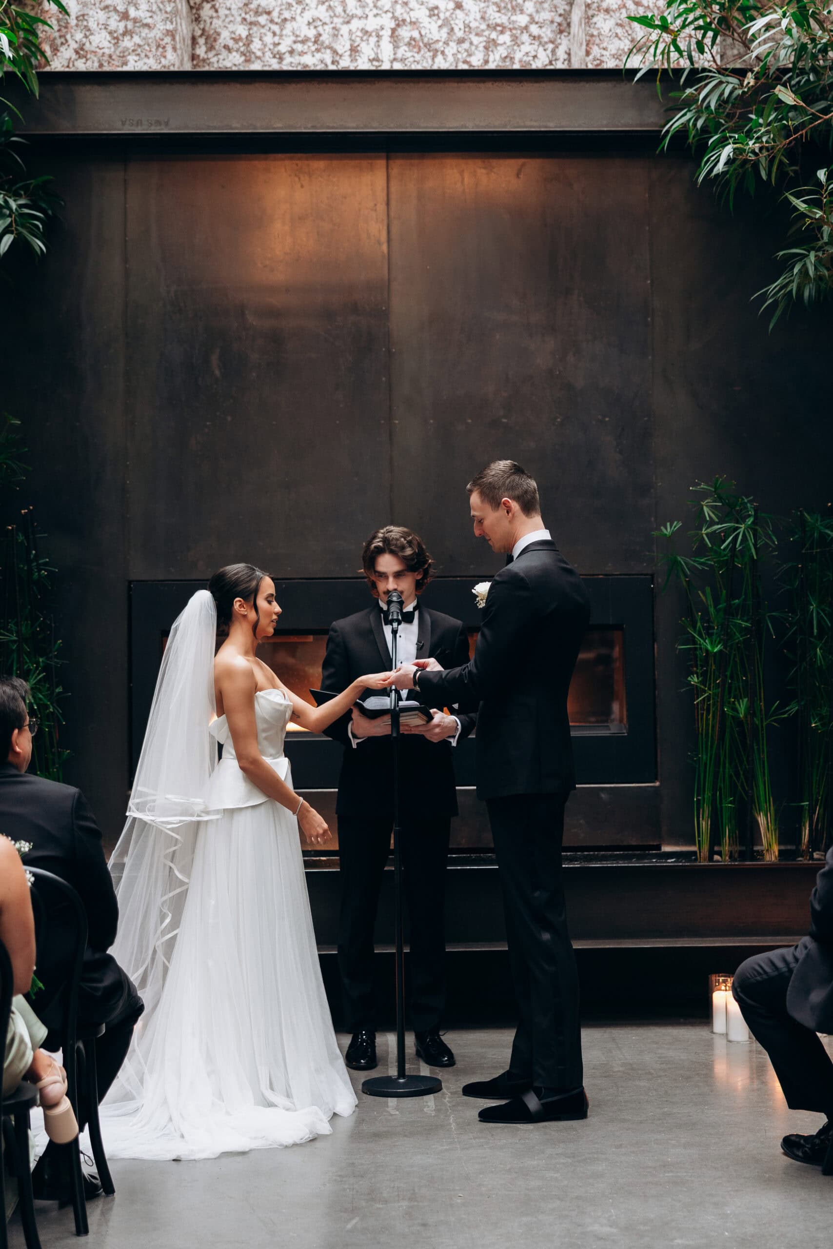 Bride and groom exchange rings during an indoor ceremony at The Foundry, standing before a dark fireplace with greenery, candles, and guests seated nearby.