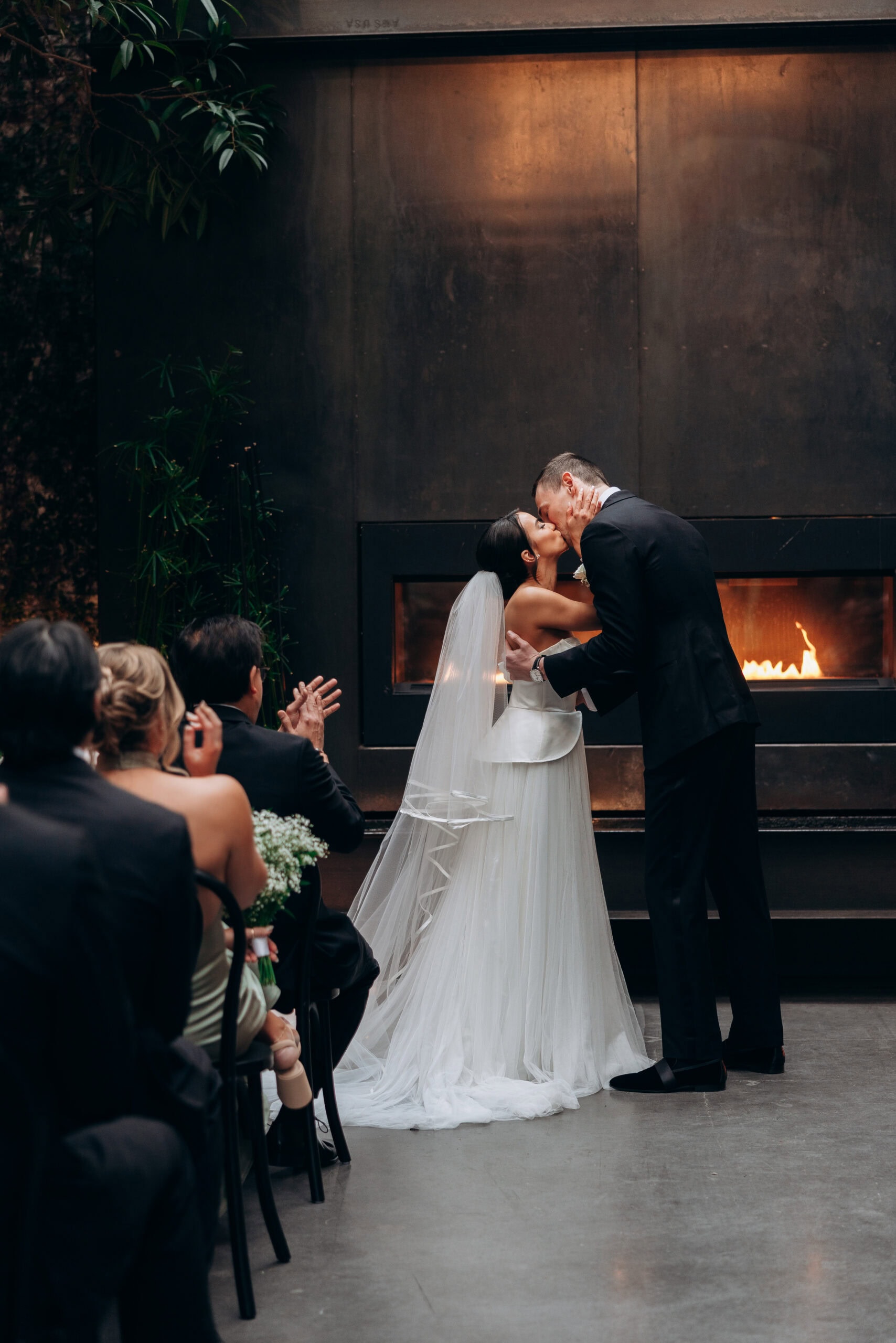 Bride and groom share their first kiss during an indoor ceremony at The Foundry, standing before a modern fireplace as seated guests applaud.
