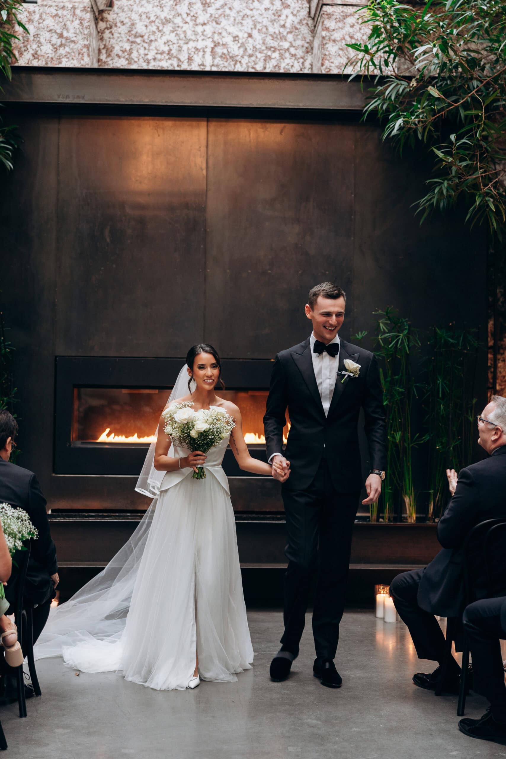 Bride and groom walk hand in hand down the aisle during an indoor wedding ceremony at The Foundry, with a glowing fireplace, candles, greenery, and seated guests.