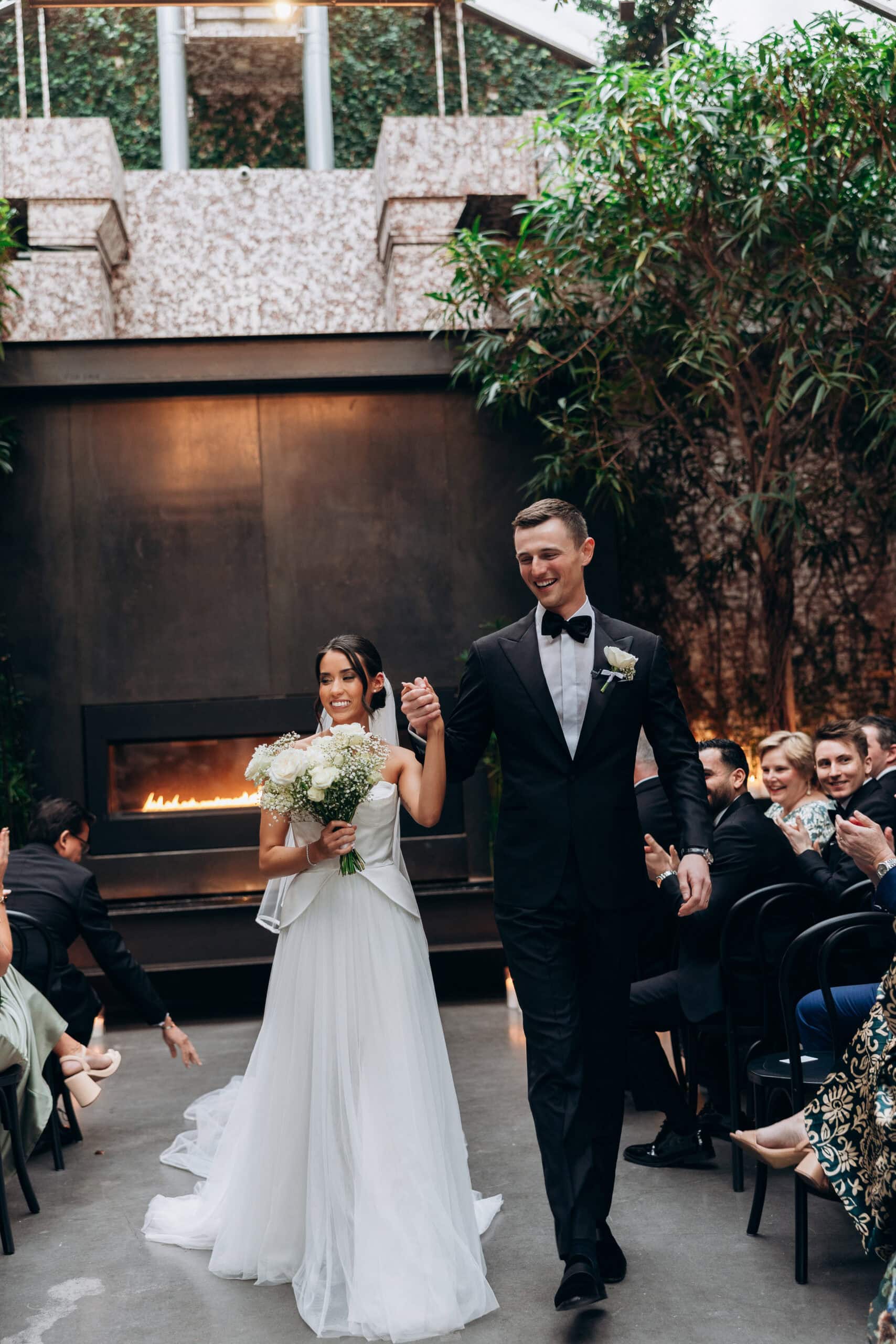 Bride and groom walk down the aisle after the ceremony at The Foundry, surrounded by seated guests, a modern fireplace, and lush indoor greenery.