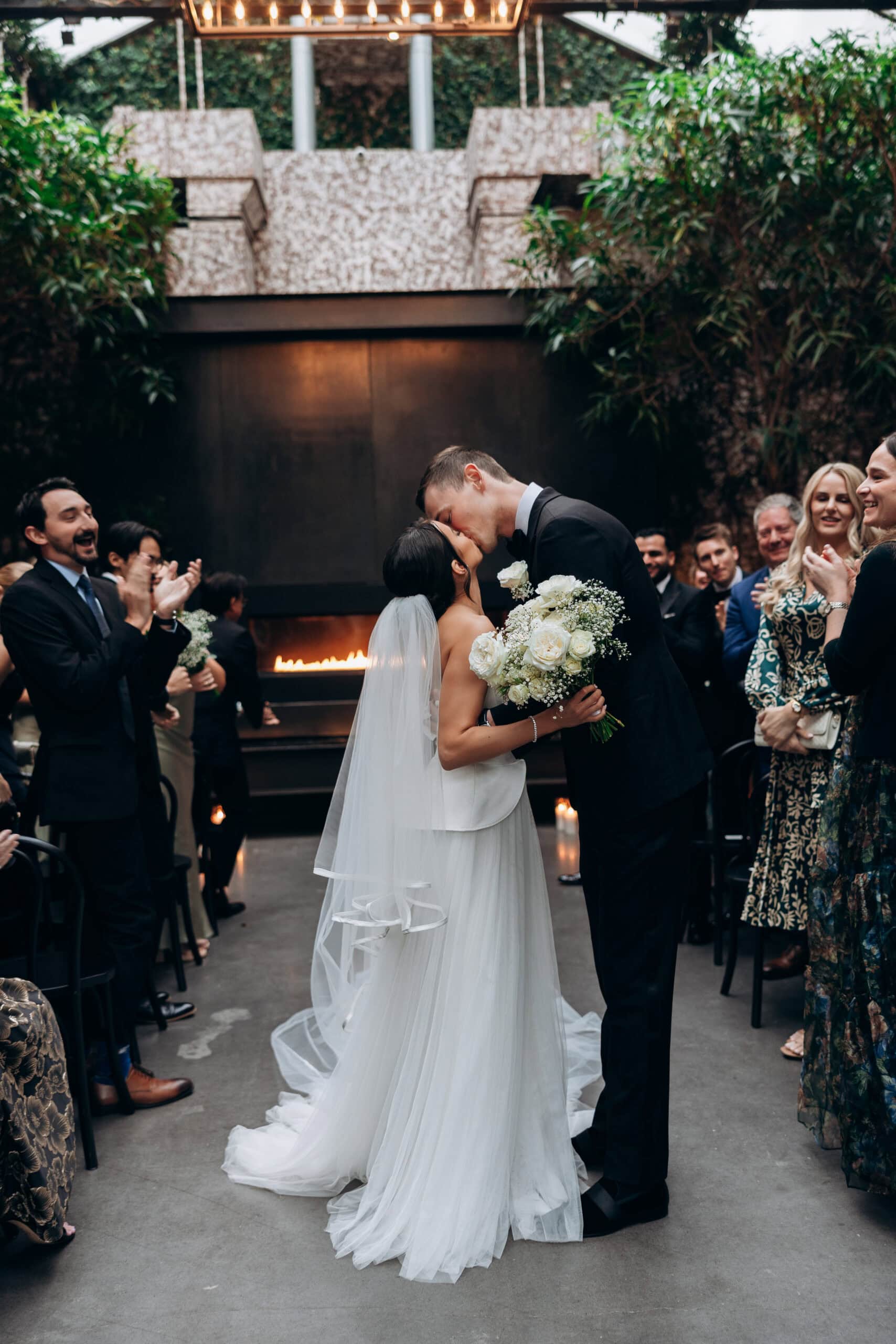 Bride and groom share their first kiss during an indoor wedding ceremony at The Foundry, framed by guests, greenery, candlelight, and a modern fireplace.