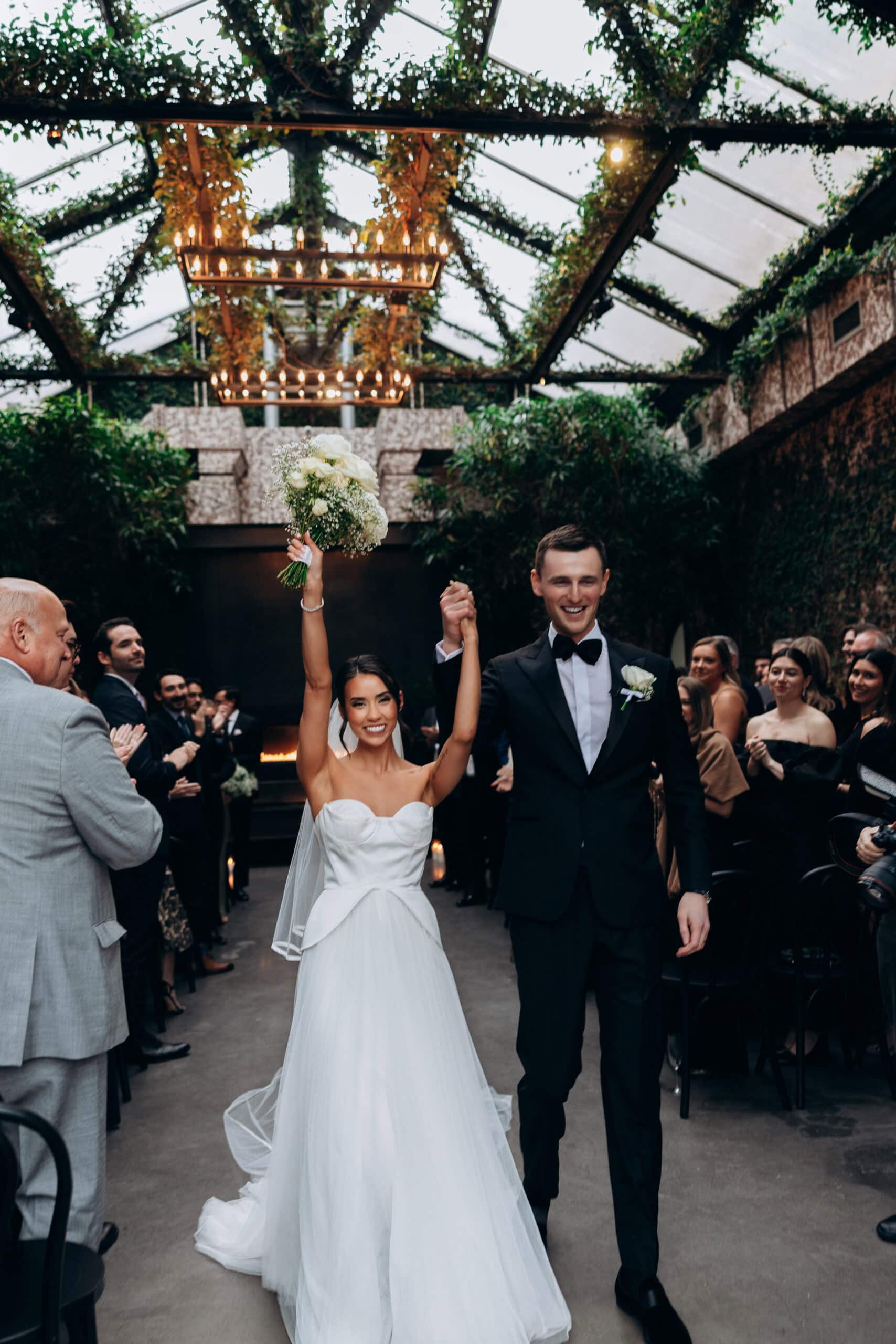 Bride and groom walk down the aisle at The Foundry after the ceremony, holding hands as the bride lifts a white bouquet beneath a glass roof with greenery and chandeliers.