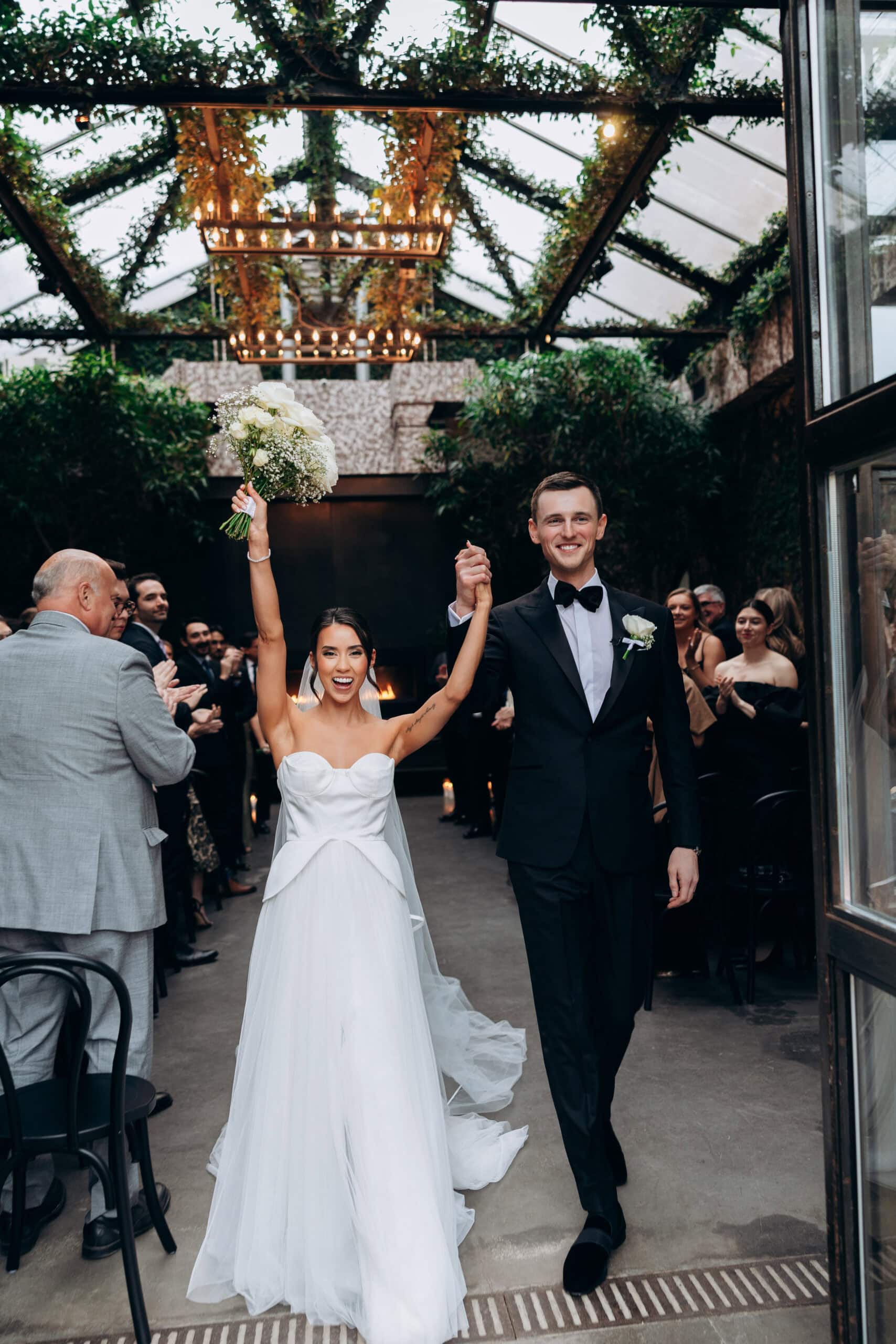 Bride and groom walk down the aisle after the ceremony at The Foundry, holding hands as the bride lifts a white bouquet beneath a glass ceiling with greenery and chandeliers while guests applaud.
