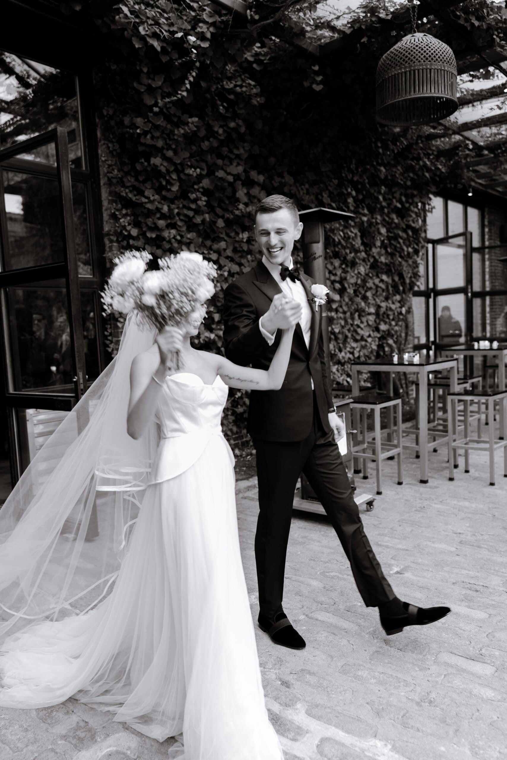 Black-and-white wedding portrait of a bride in a strapless gown and long veil holding bouquets beside a groom in a tuxedo in the ivy-covered courtyard at The Foundry.