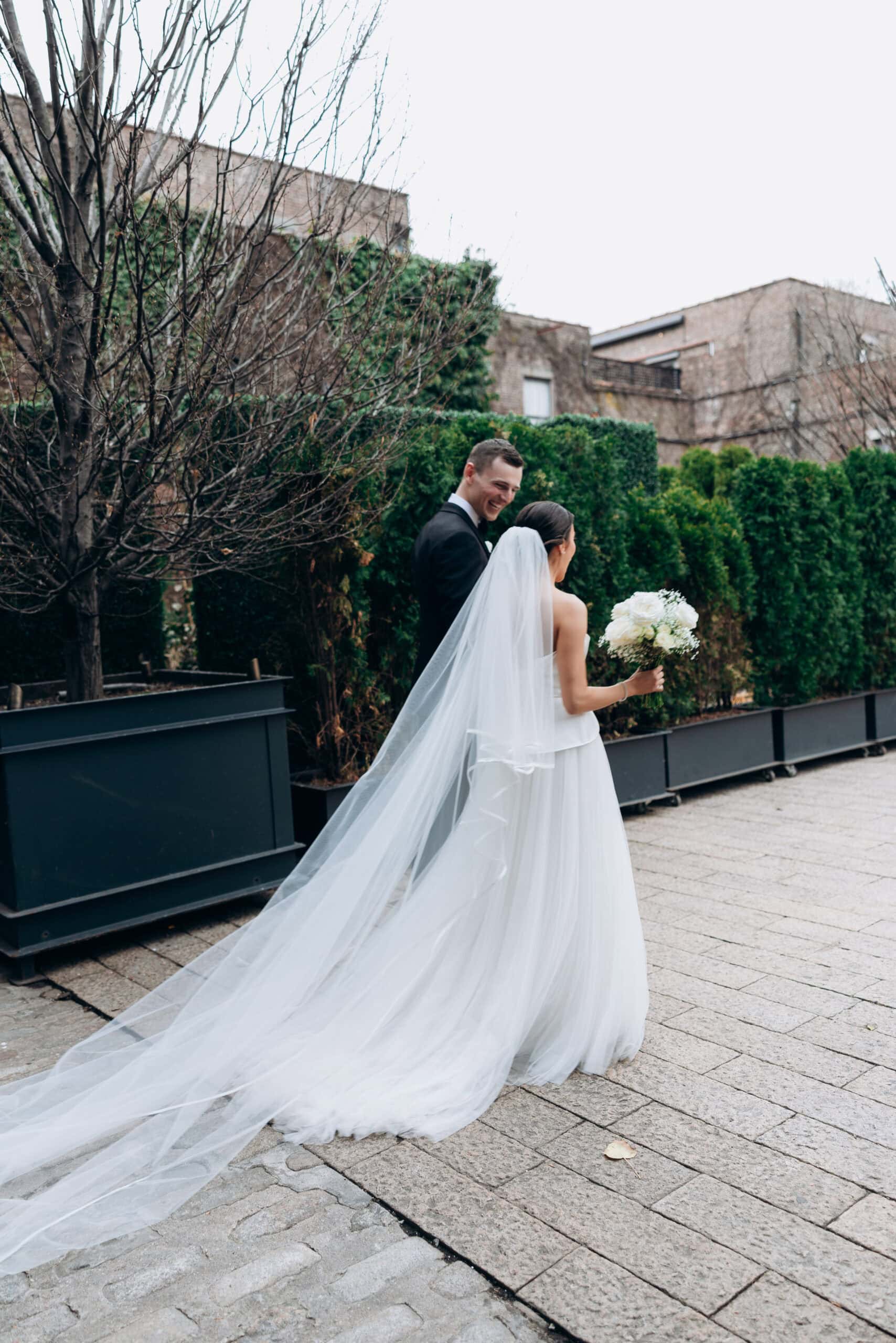 Bride with a long veil and white bouquet walks with her partner in a courtyard at The Foundry, framed by hedges, bare trees, and brick buildings.
