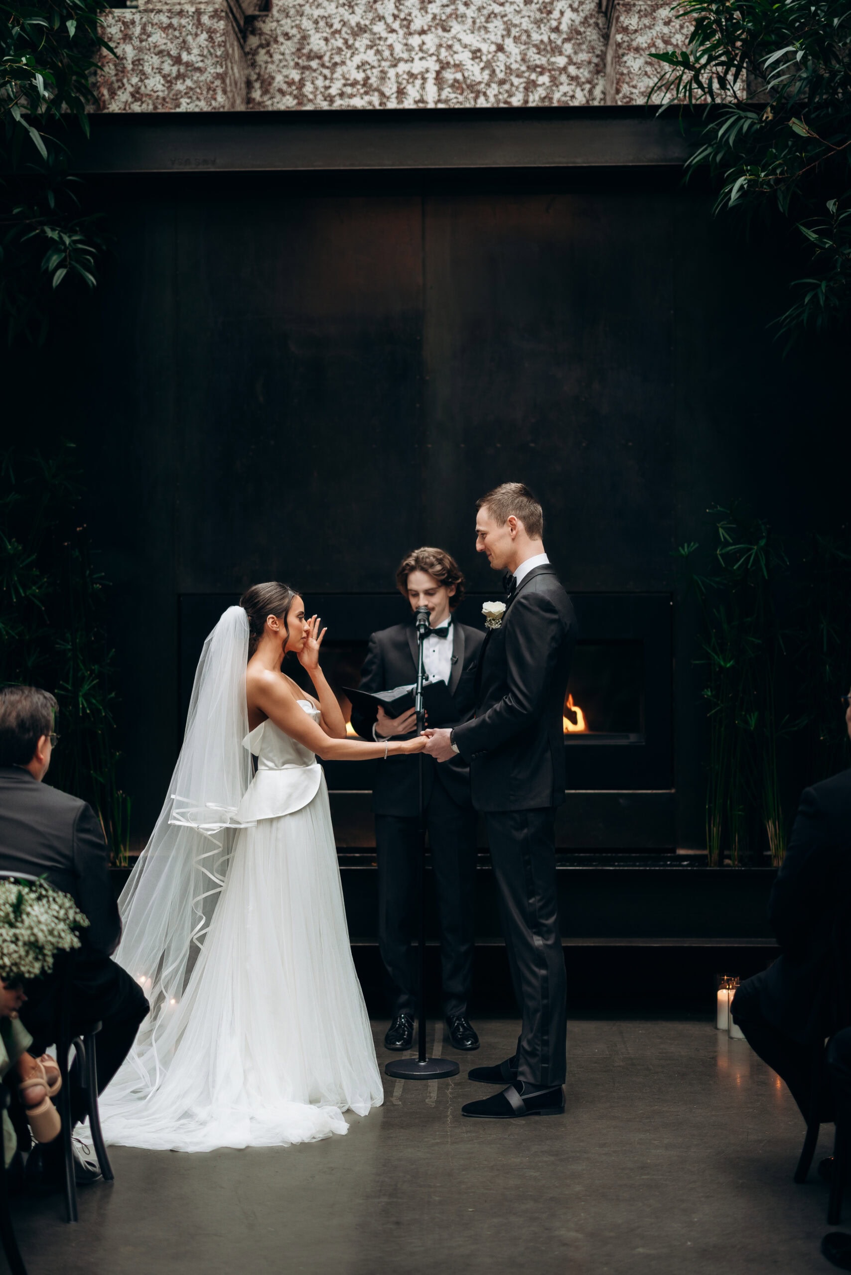 Bride and groom exchange vows during an indoor ceremony at The Foundry, standing before a dark fireplace with an officiant, candles, and tropical greenery.