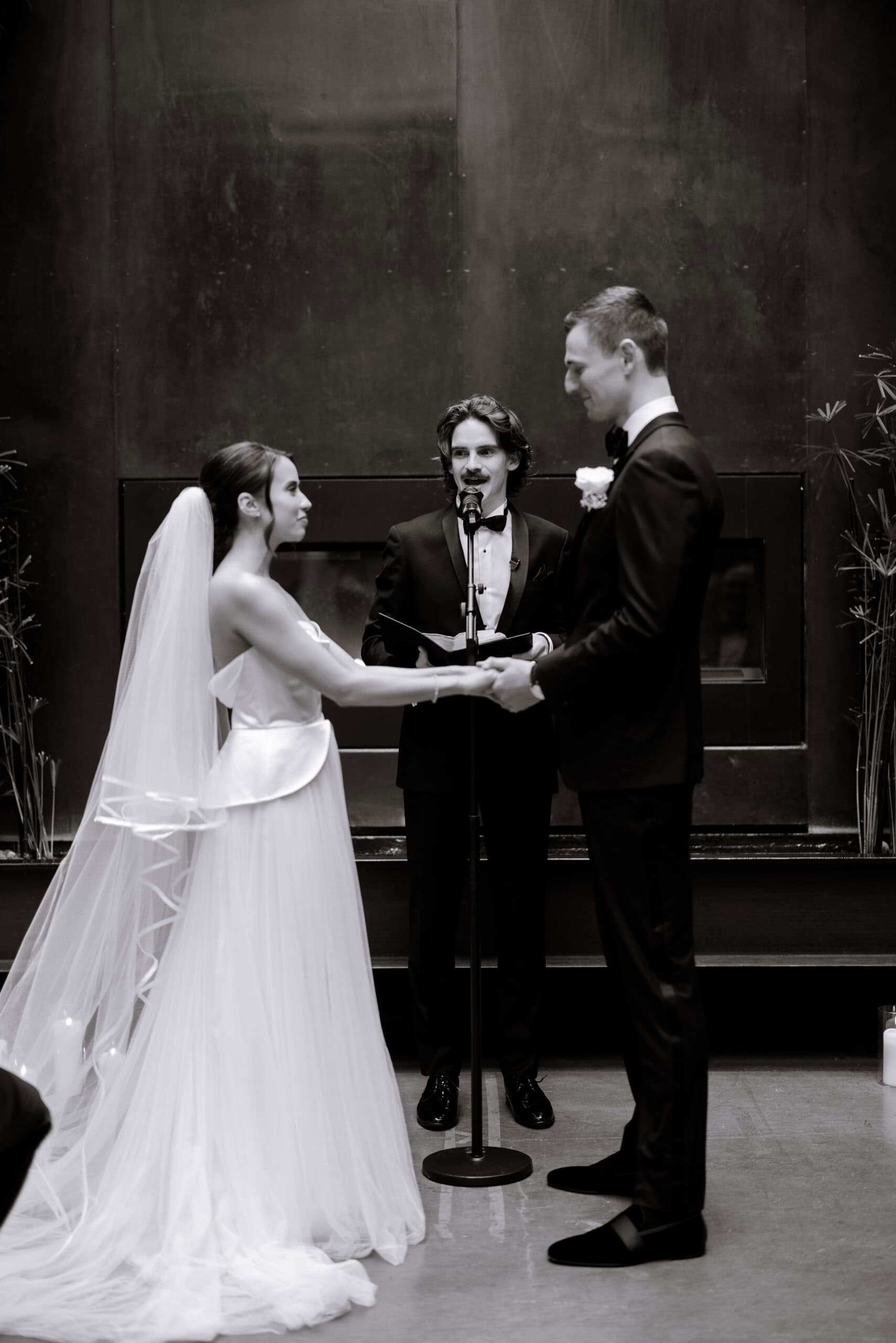 Black-and-white wedding ceremony at The Foundry with the couple holding hands before the officiant, framed by a dark fireplace wall and a long veil.