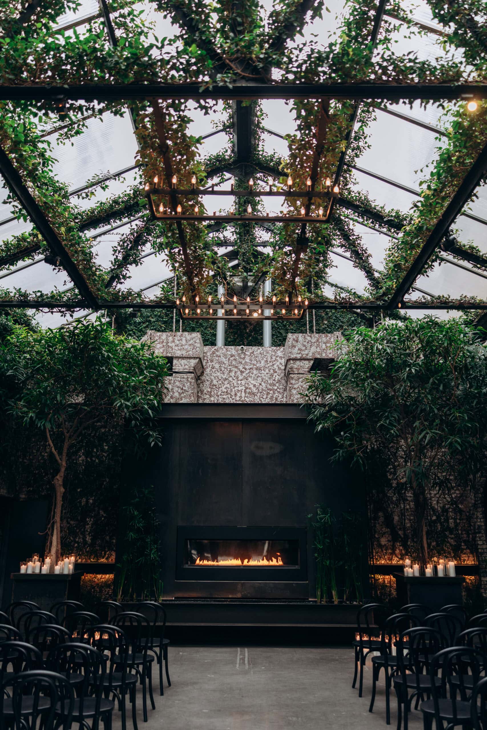 Indoor ceremony setup at The Foundry with a glass ceiling, hanging greenery, chandeliers, black chairs, candles, and a central fireplace.