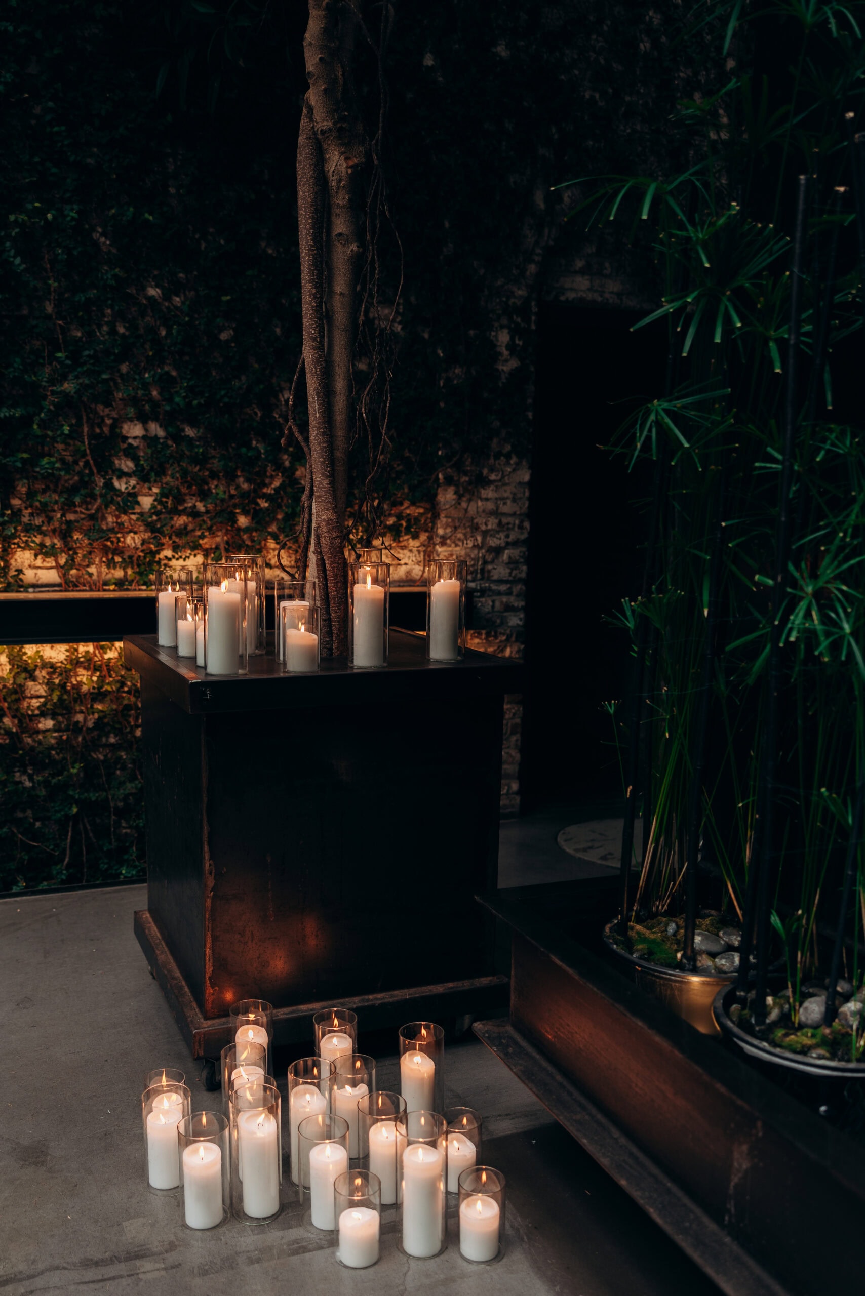Candlelit courtyard detail at The Foundry with glass votives around a sculptural tree, dark greenery, and textured stone walls in low evening light.