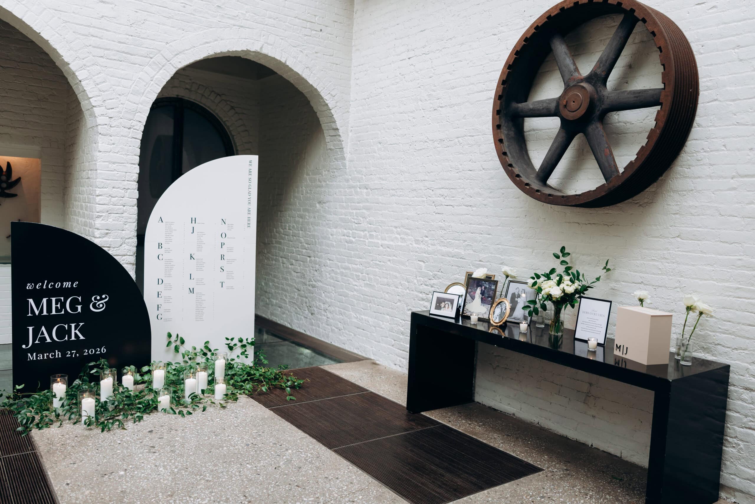 Wedding welcome display at The Foundry with black and white signage for Meg and Jack, candles, greenery, framed photos, and white brick arches beneath a large industrial wheel.