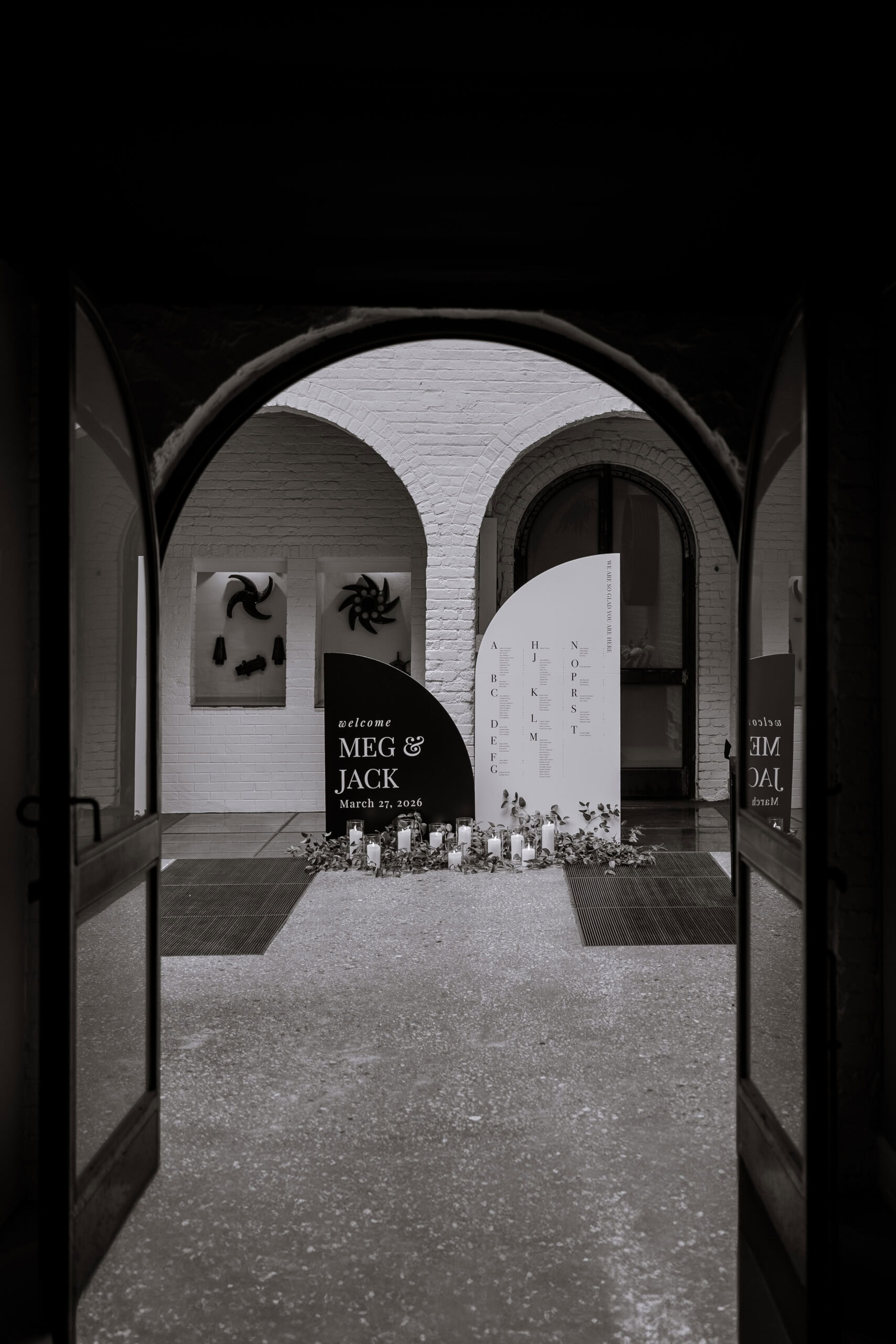 Black-and-white view through open doors of a wedding welcome display at The Foundry, with curved signs for Meg and Jack, candles, greenery, and arched brick walls.