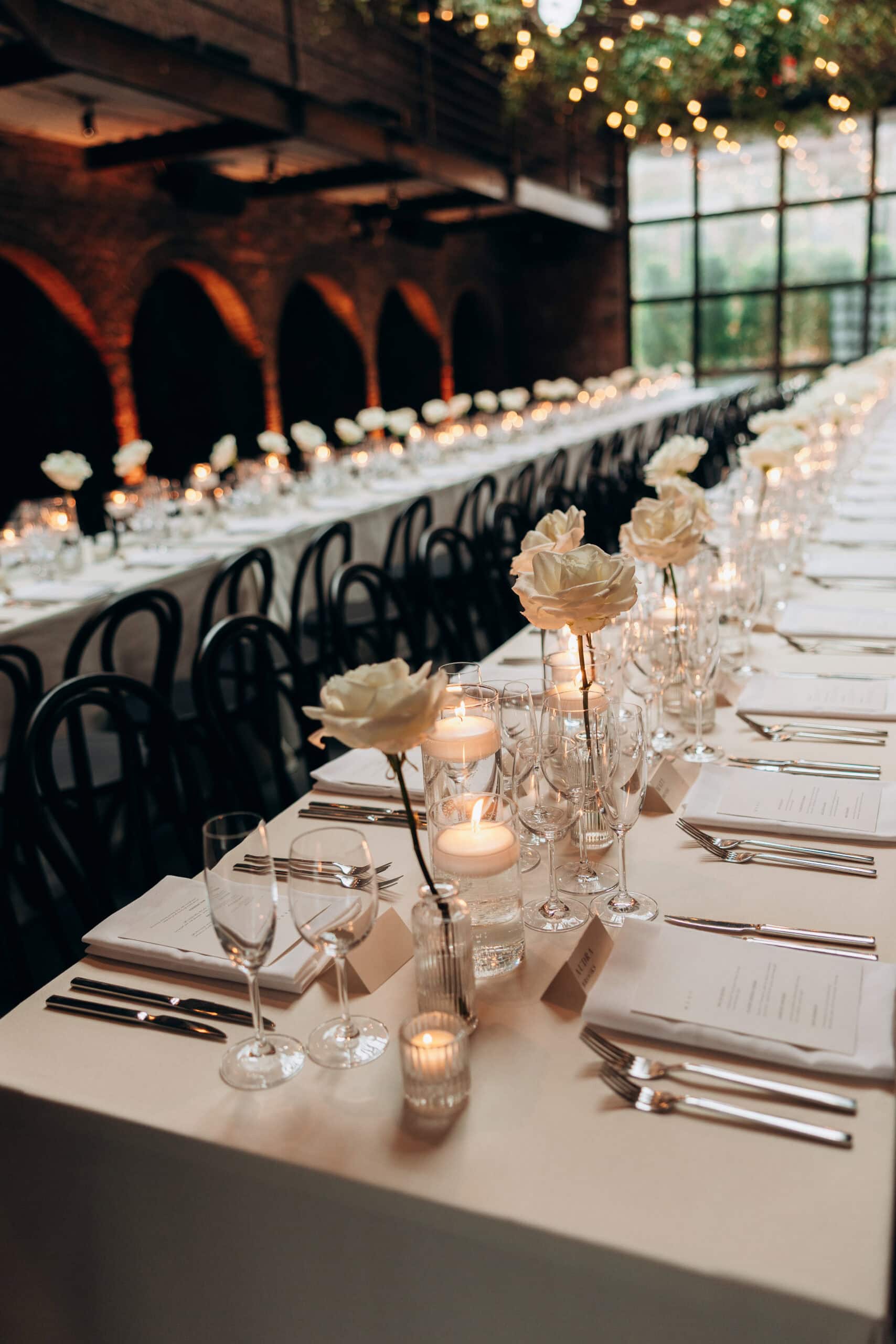 Long candlelit wedding reception table at The Foundry with ivory roses, black chairs, fine glassware, white linens, brick arches, and large industrial windows.