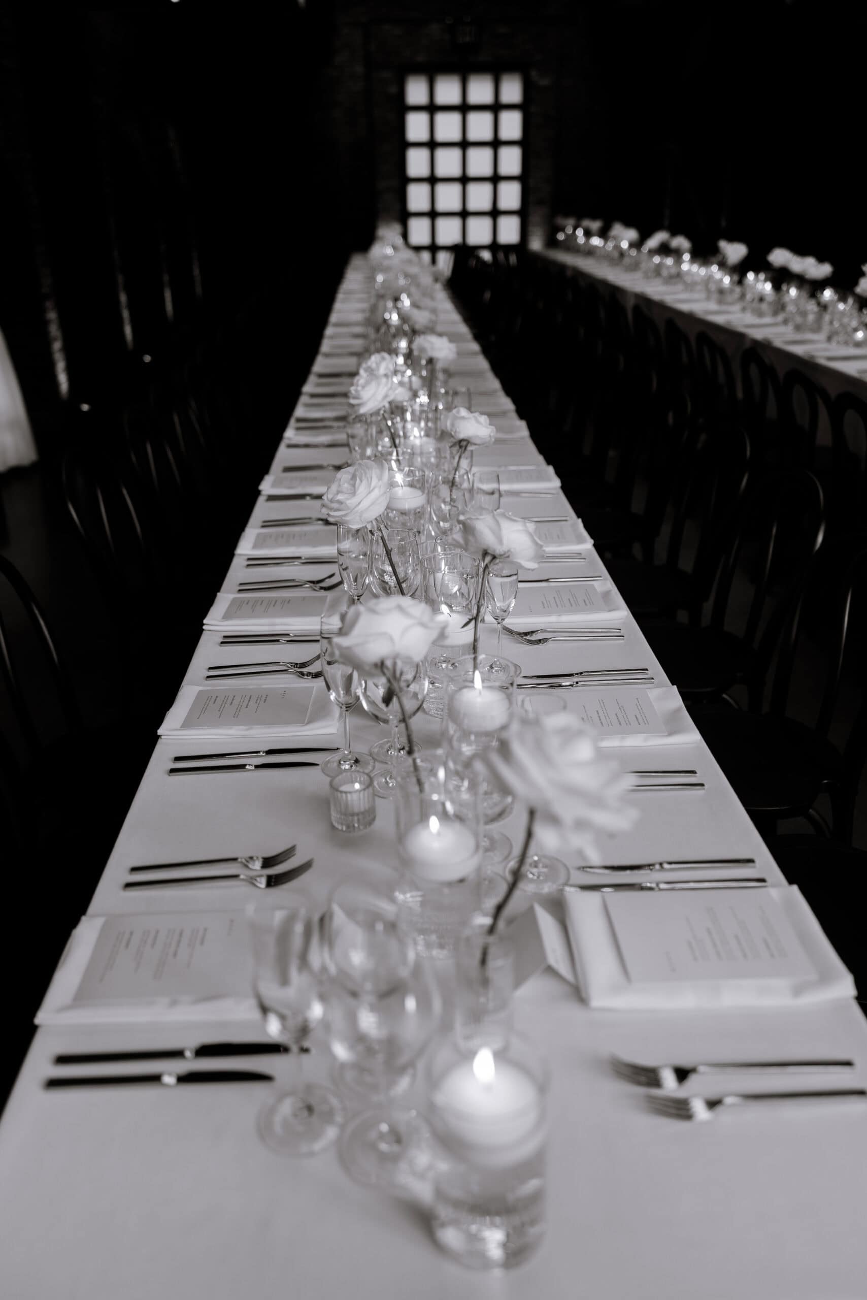 Black-and-white wedding reception setup at The Foundry with a long table, white roses, candles, menus, and glassware leading toward a glowing window.