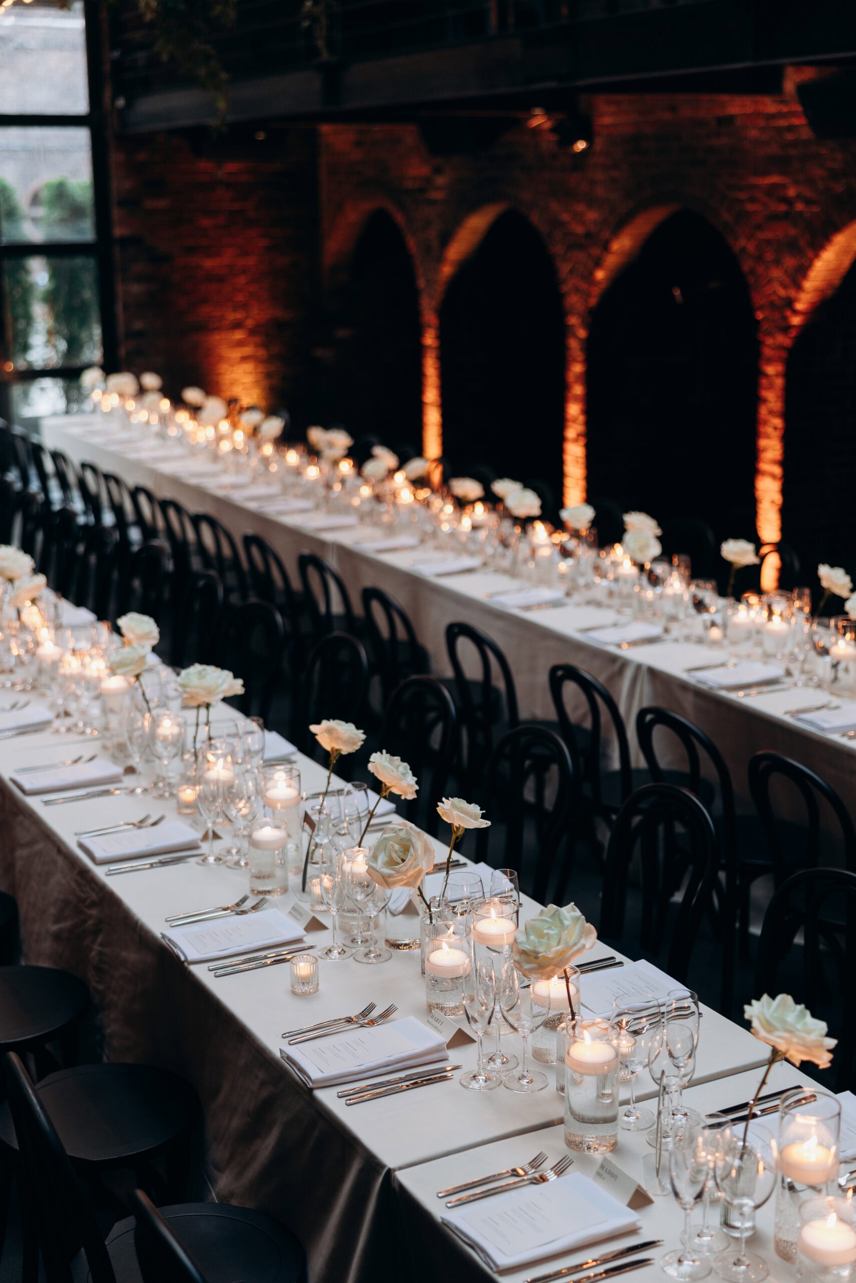 Candlelit wedding reception at The Foundry with long white-clothed tables, black chairs, white roses, glassware, and brick arches glowing with amber uplighting.
