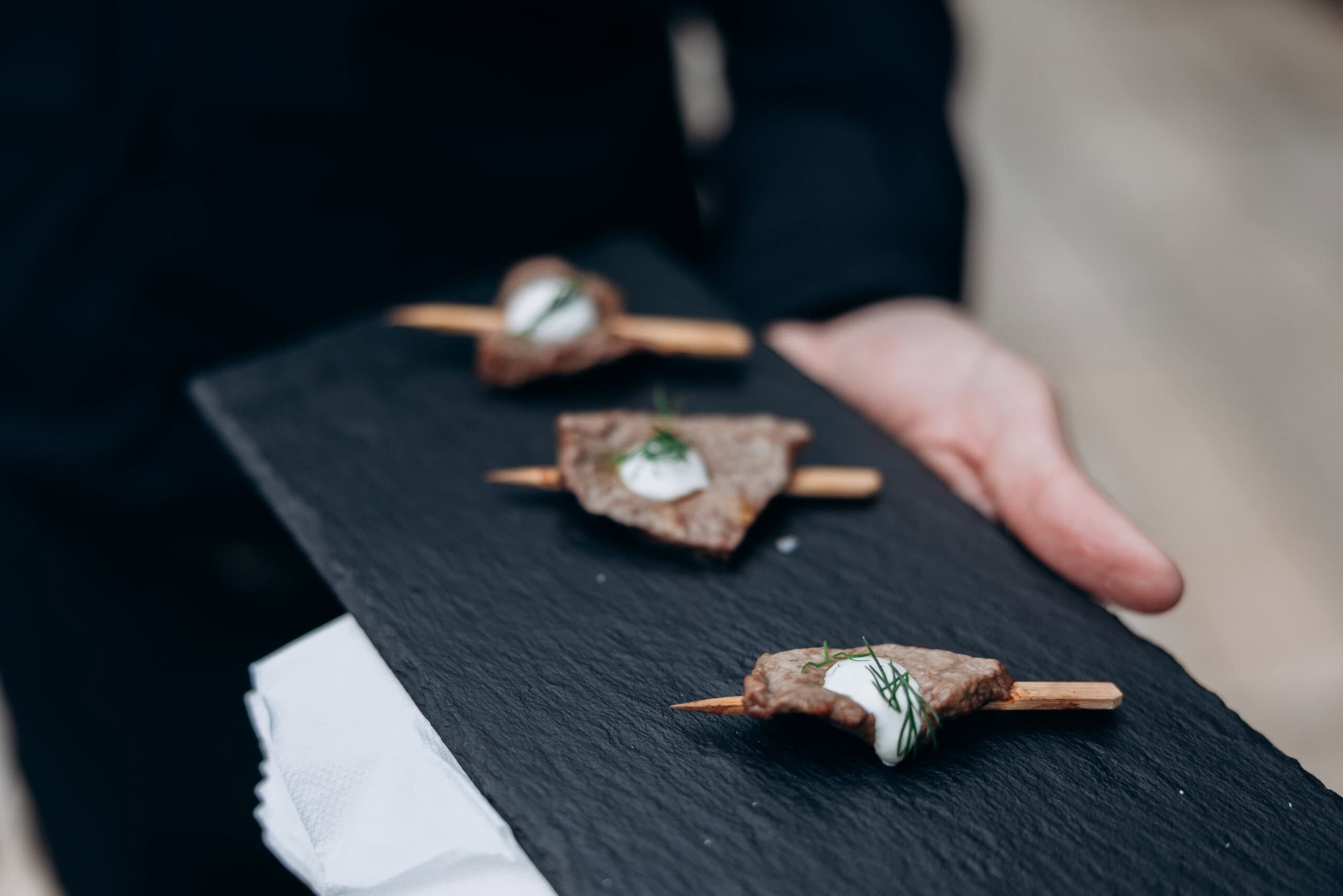 Close-up of a server holding a black slate tray with three skewered hors d’oeuvres topped with white sauce and herbs during a wedding reception at The Foundry.