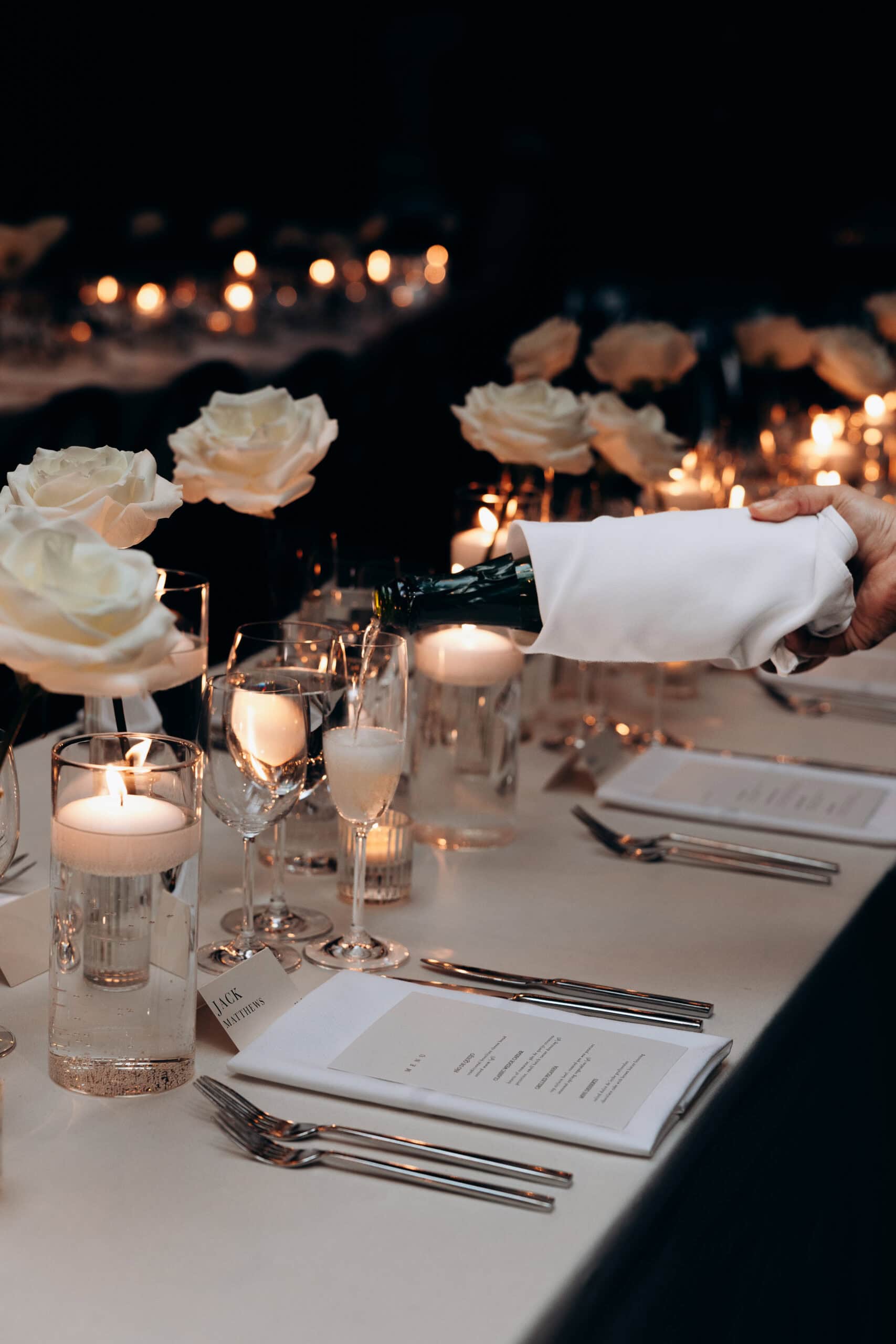 A hand pours champagne into a flute at a candlelit wedding reception table at The Foundry, styled with white roses, votive candles, menus, and silver flatware.