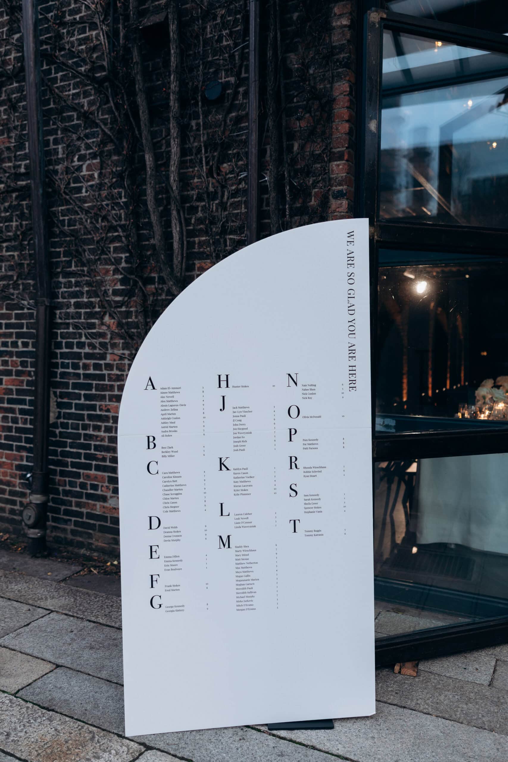 Modern white wedding seating chart with curved top outside The Foundry, set against dark brick walls, bare vines, and a glass entrance.