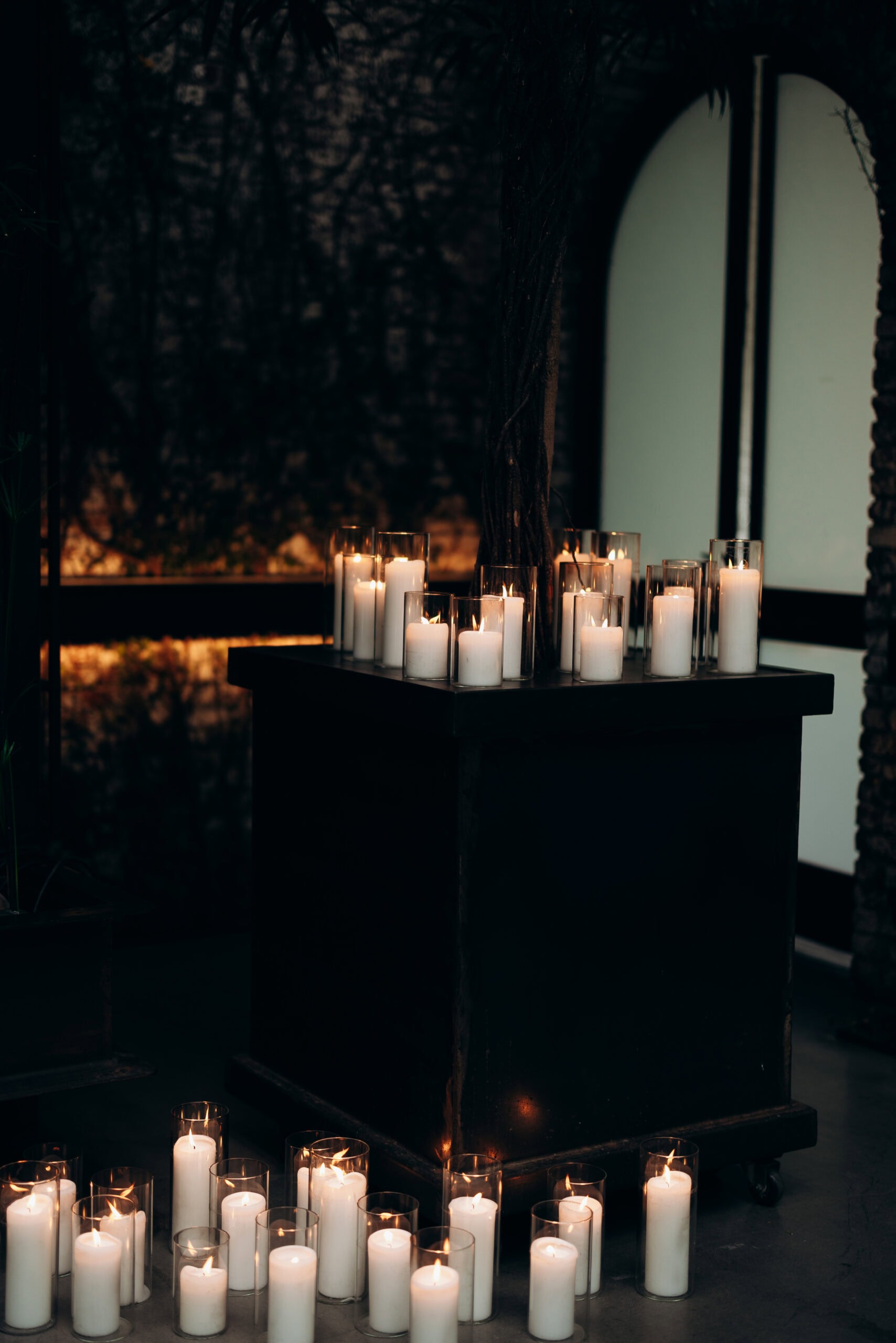 Dozens of white pillar candles in clear glass cylinders surround a dark pedestal indoors at The Foundry, creating a moody, candlelit wedding ambiance.