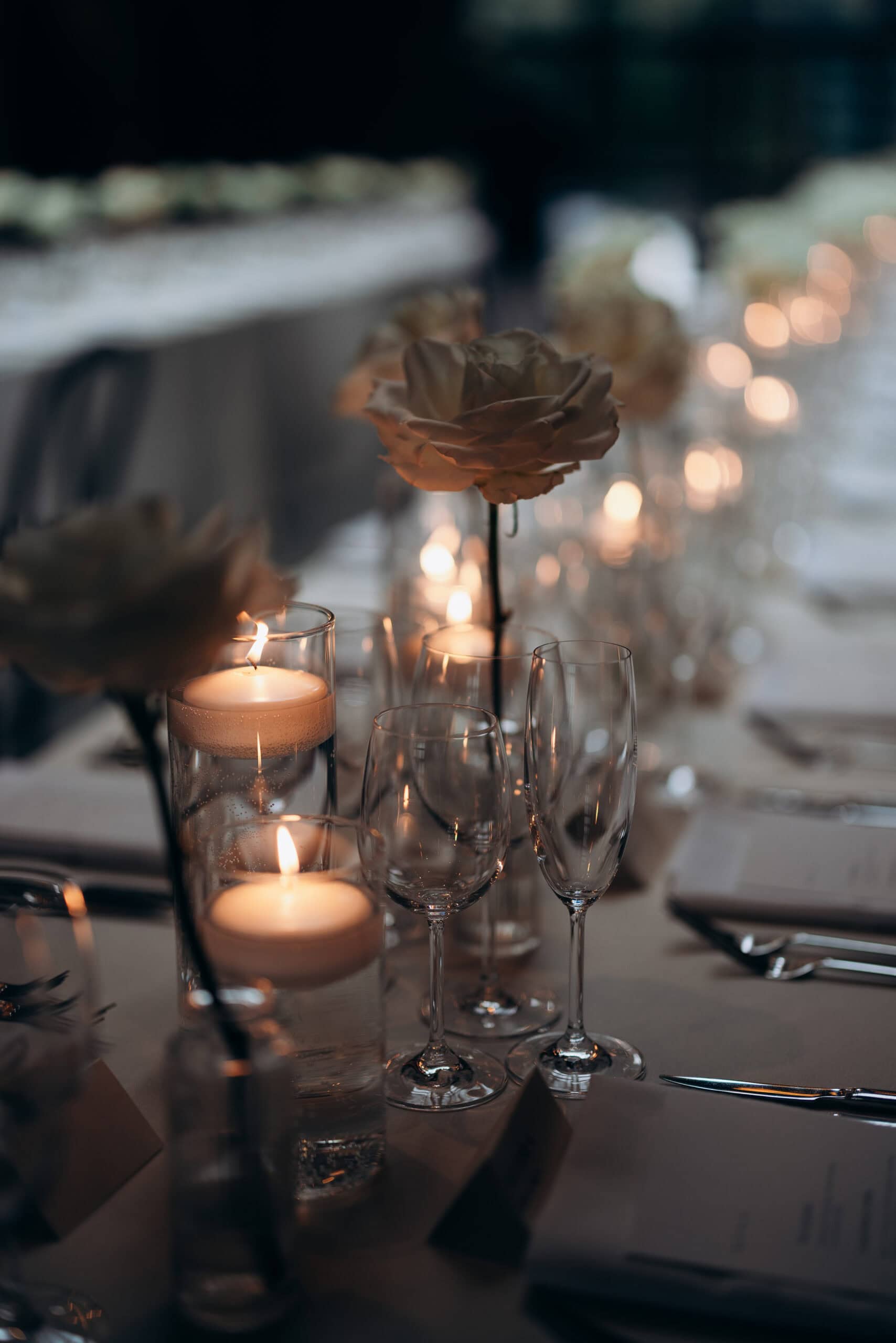 Close-up of a candlelit wedding reception table at The Foundry with ivory rose centerpieces, clear stemware, and soft evening bokeh in a refined indoor setting.