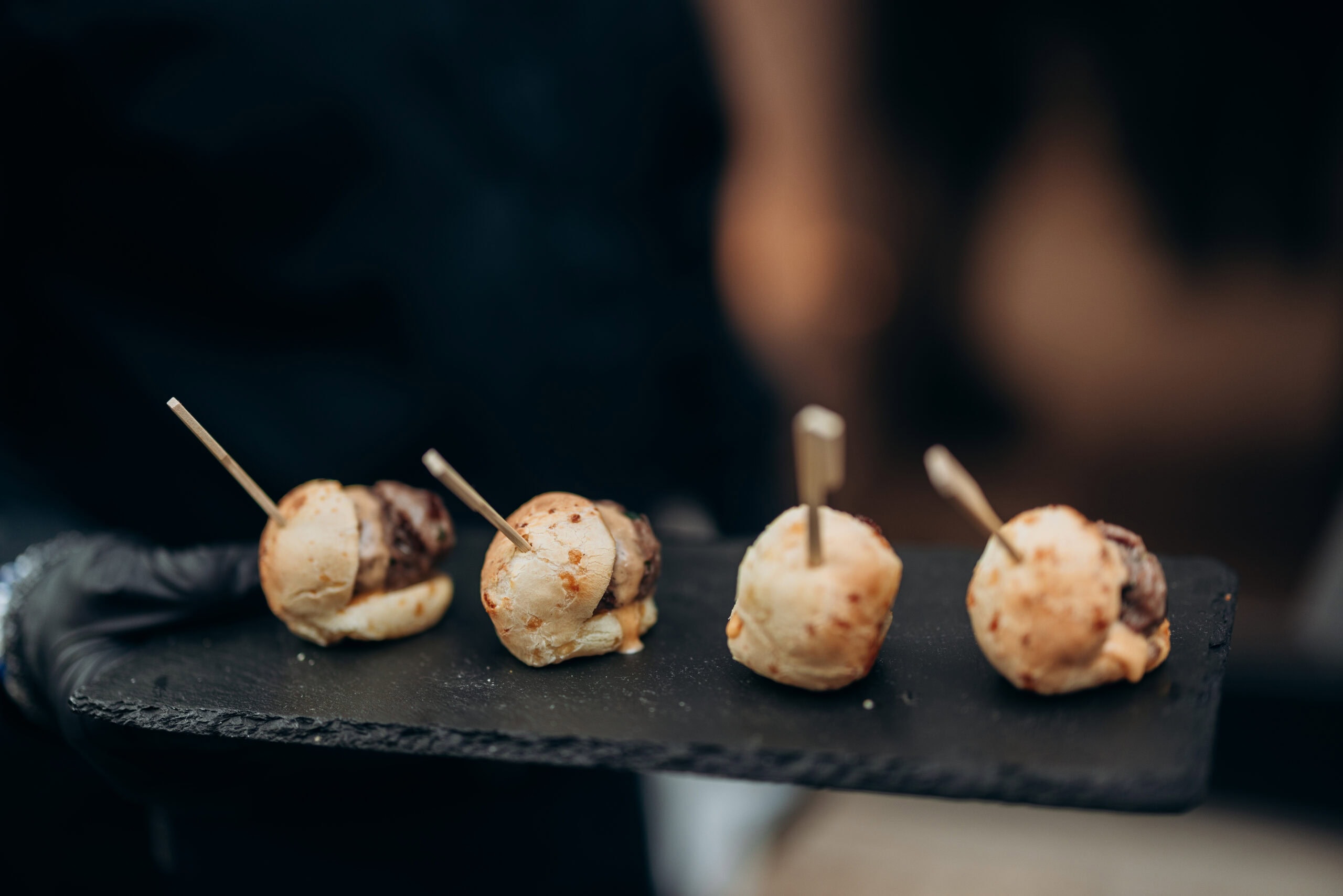 Close-up of four miniature sliders on a black slate serving tray, held by a gloved server during a wedding reception at The Foundry.