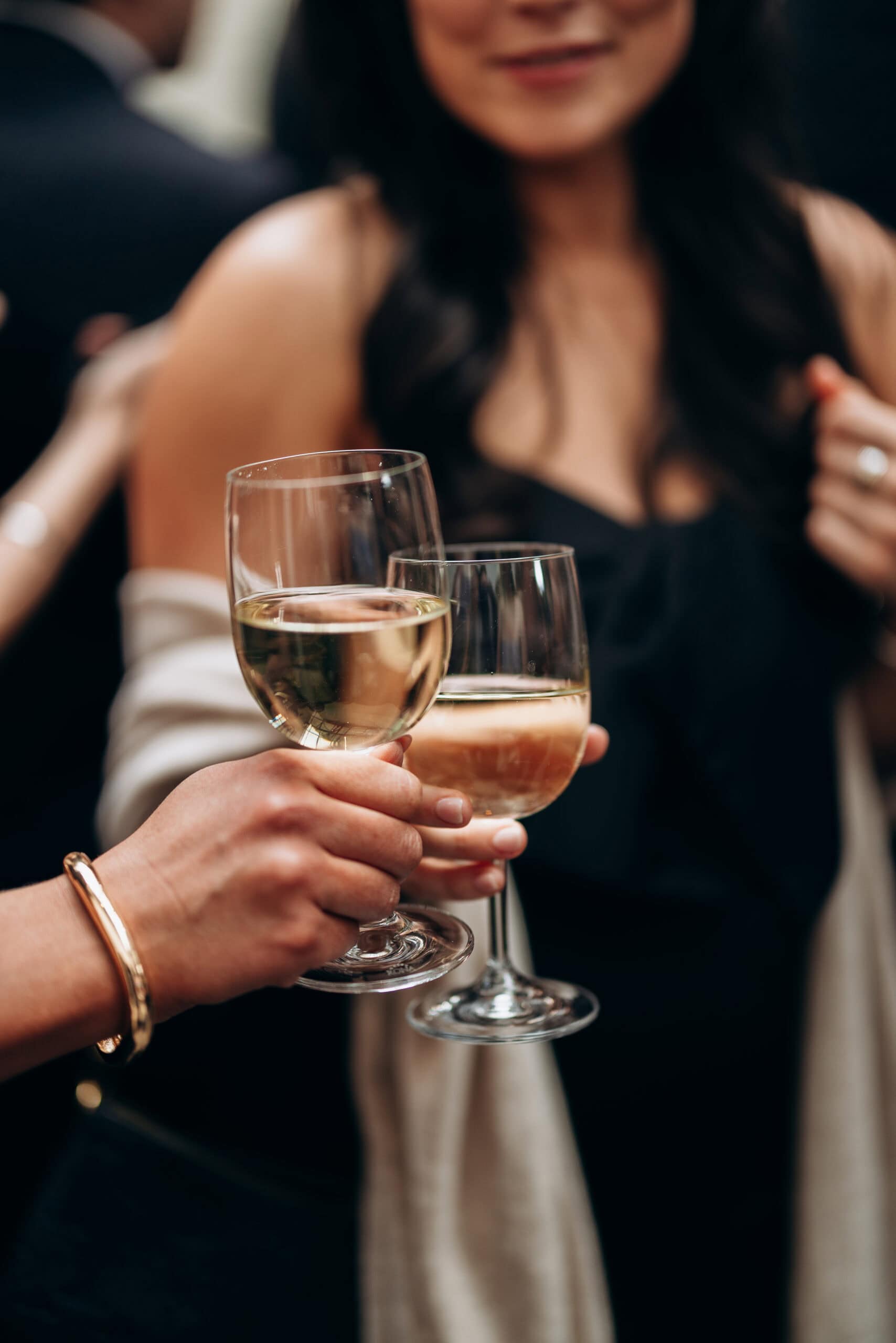 Close-up of two guests holding glasses of white wine during a wedding reception toast at The Foundry, with softly blurred figures in dark formal attire behind them.