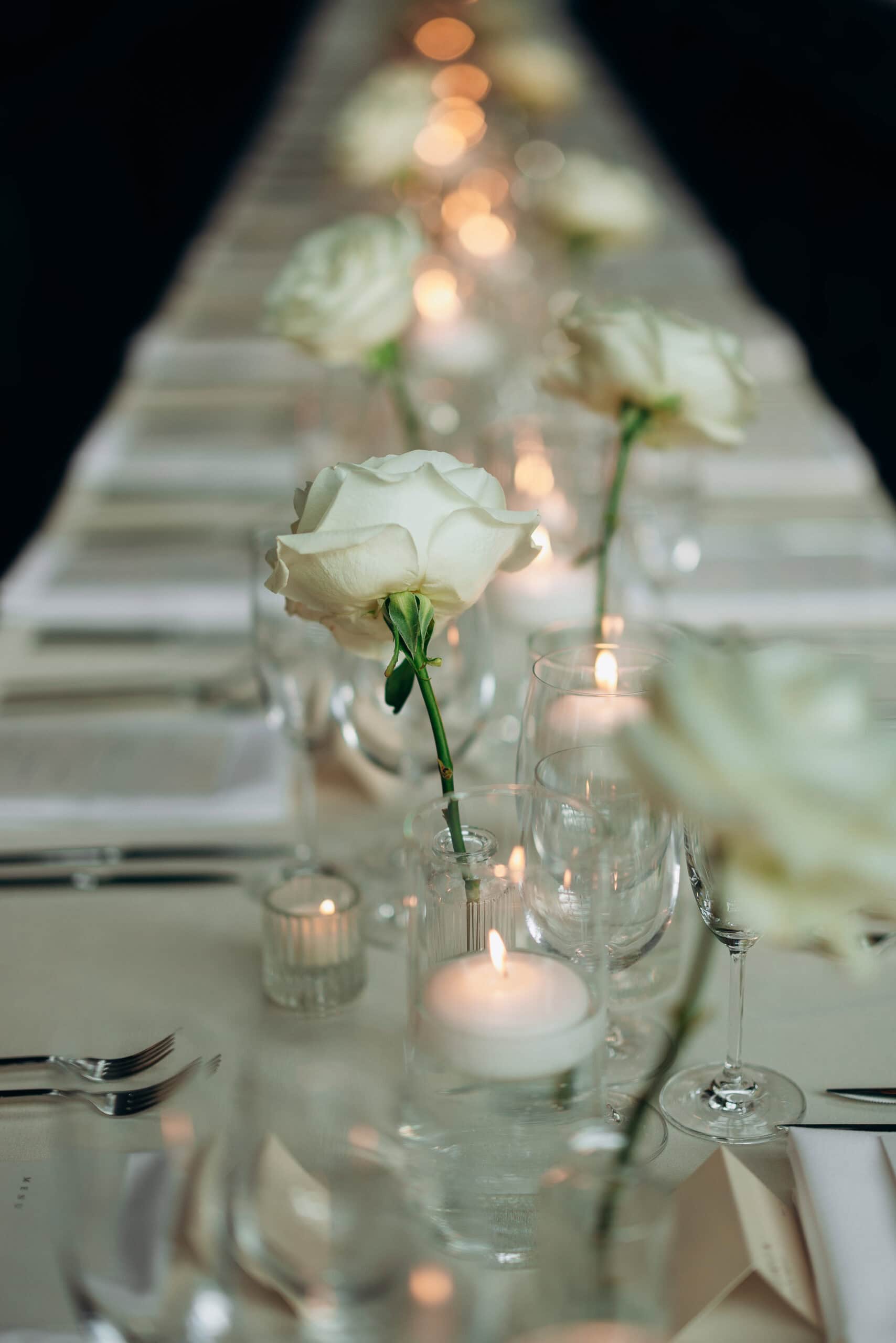 Reception table at The Foundry with white roses in clear vases, lit candles, folded napkins, and glassware arranged in a long, softly blurred row.