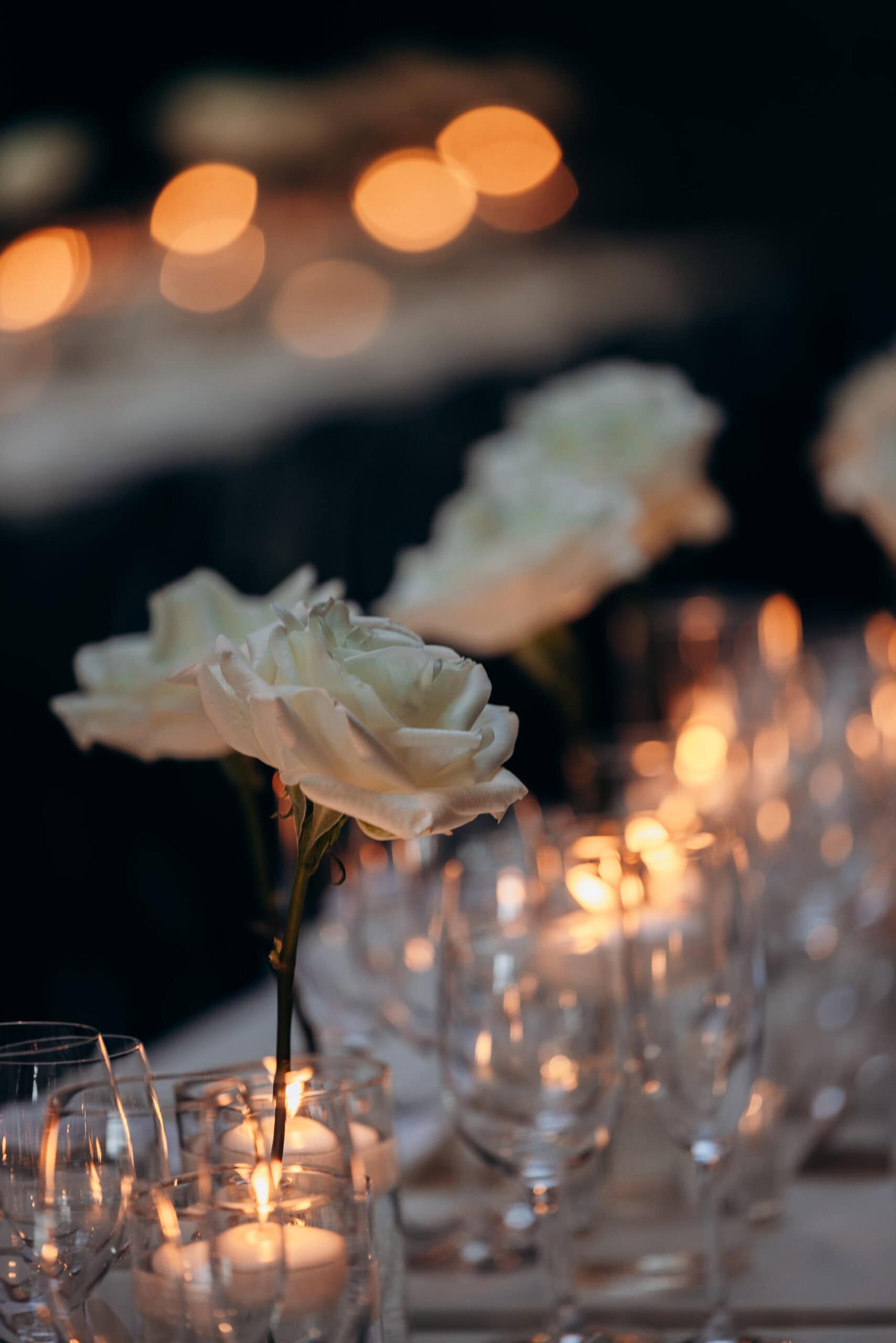 White roses in clear glass vases with candlelight and stemware on a dim reception table at The Foundry.