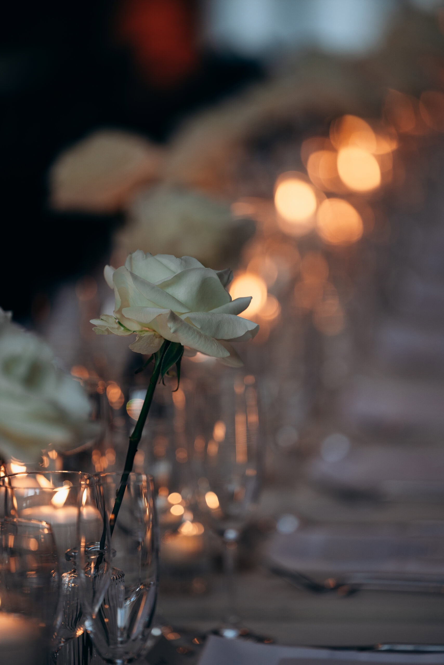 Close-up of an ivory rose in a crystal vase on a candlelit reception table at The Foundry, with soft amber bokeh, glassware, and place settings fading into blur.
