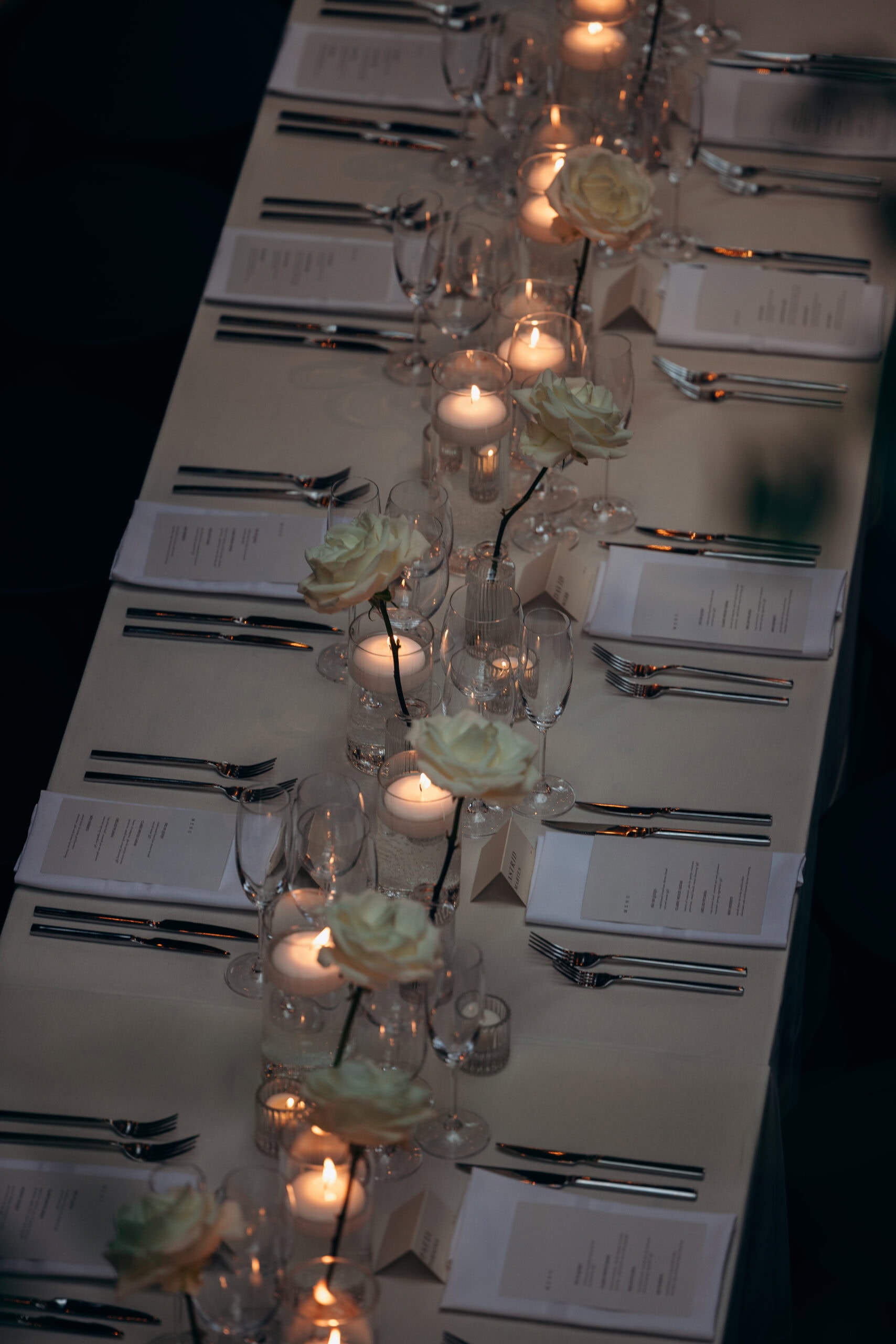 Candlelit wedding reception table at The Foundry with ivory roses in bud vases, white menus, folded napkins, silver flatware, and clear glassware arranged in a long row.
