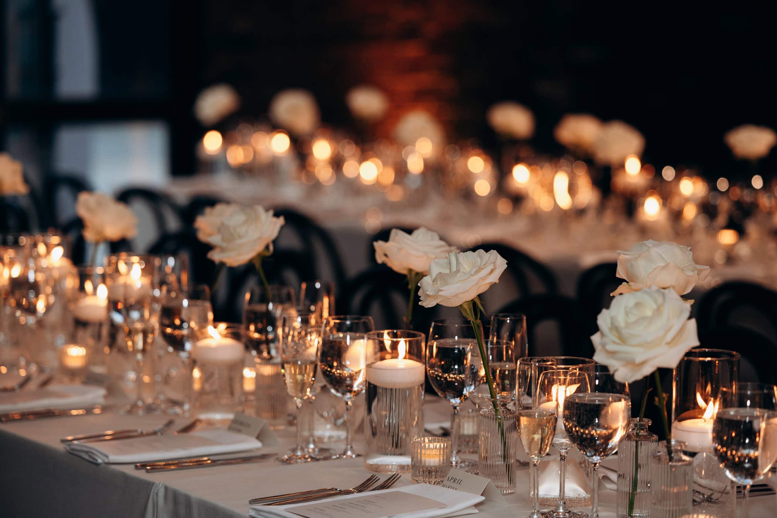Candlelit wedding reception tables at The Foundry with white roses, glassware, folded menus, and glowing votives in a dim, elegant interior.