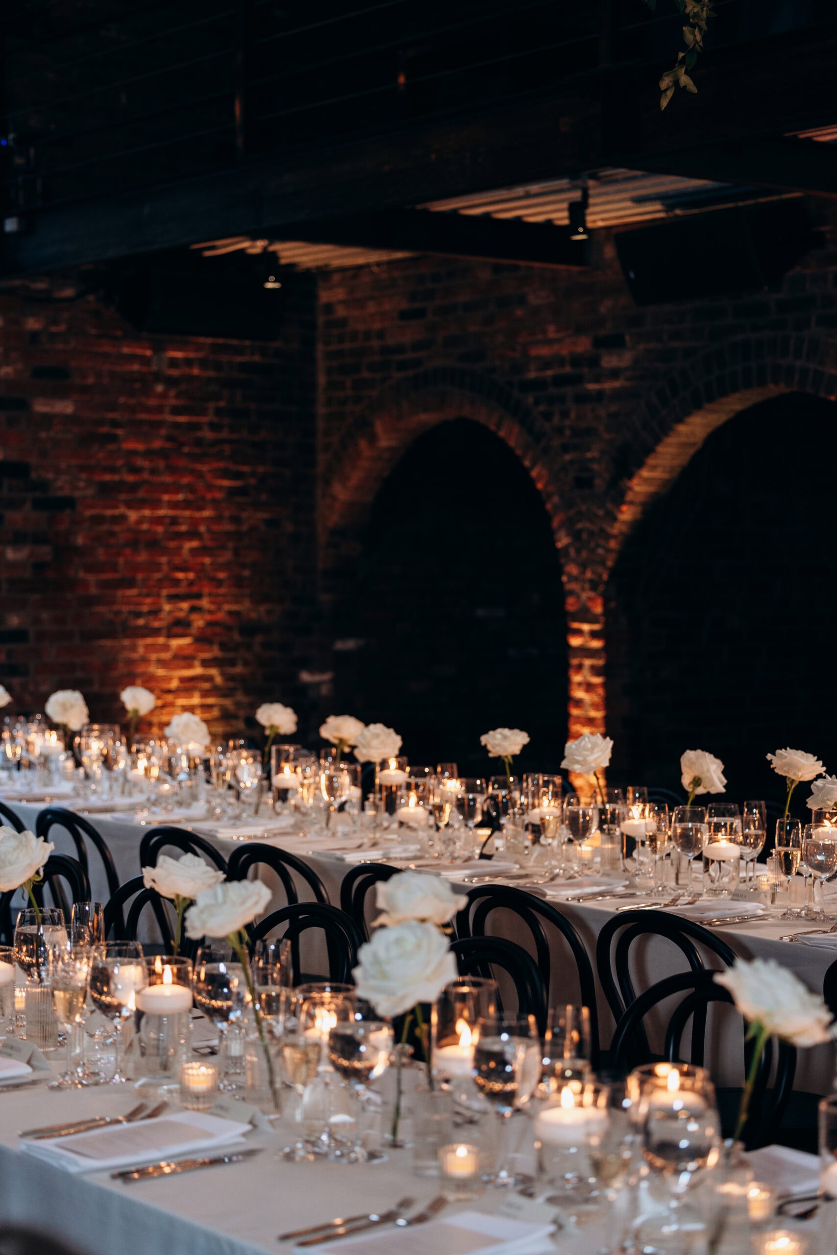 Long candlelit reception tables with white roses, glassware, and black bentwood chairs beneath brick arches at The Foundry.